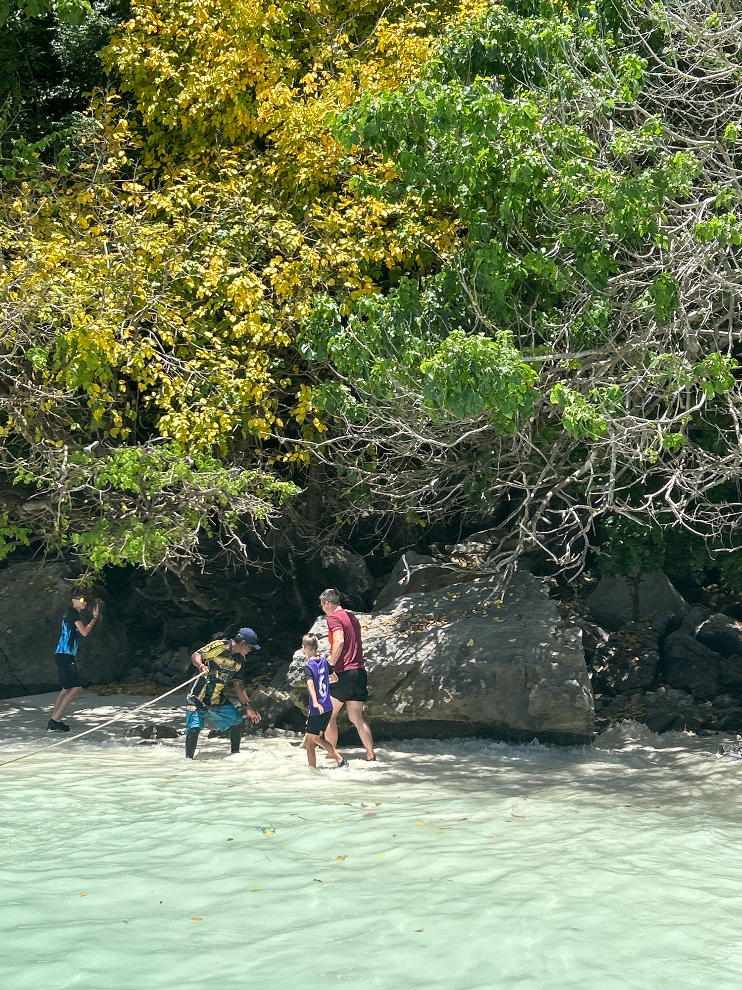 In the shallow, clear, turquoise waters of Ton Sai Beach, Thailand, four individuals are present. In the foreground, a person wearing a blue and yellow life vest and a dark blue baseball cap is holding a rope taut. To their right, a person in a red t-shirt and black shorts stands facing away from the camera. To the left, a person wearing a blue shirt with black shorts is walking away from the water. A young person in a purple shirt with the number '6' on it is walking towards the water. The individuals are near a rocky shoreline, partially shaded by a dense canopy of green and yellow foliage. The water exhibits a gentle wave action near the shore.