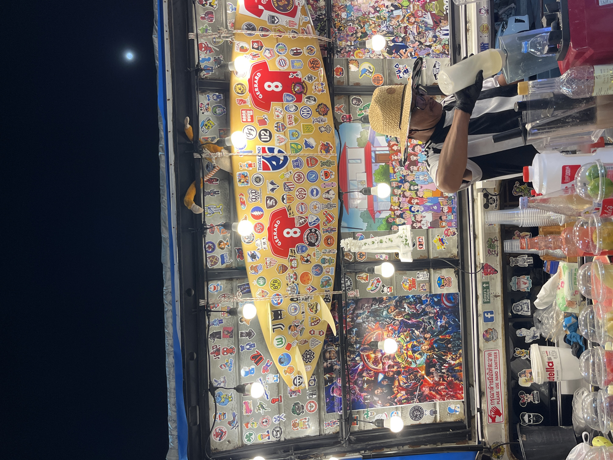 The image depicts a street food vendor in Pattaya City, Thailand at night. The vendor is seen preparing drinks, wearing a straw hat and black gloves. The stall is decorated with numerous stickers, posters, and a surfboard. A sign reads "Please Use Hand Sanitizer" in Thai and English, and a Liverpool Football Club sticker is also visible. The sky is dark with a visible moon.