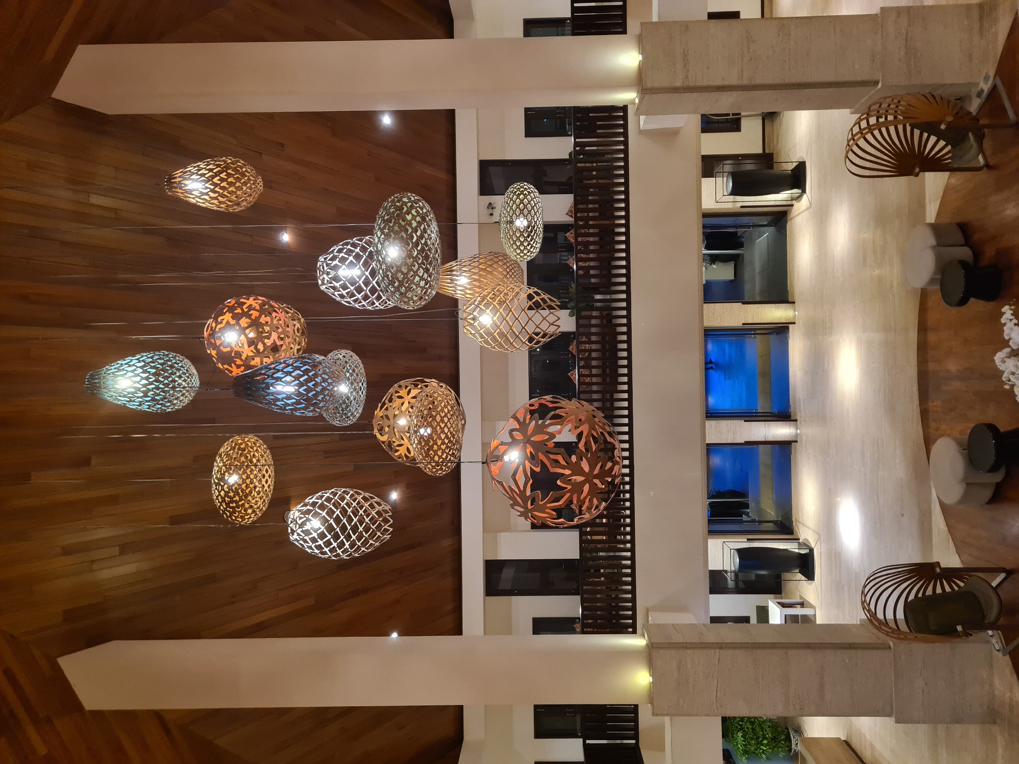 This media file depicts the interior of a modern hotel lobby in Da Nang, Vietnam. The scene is dominated by a multi-level atrium with a warm, inviting ambiance, likely illuminated by artificial lights to suggest an evening or indoor setting. A prominent feature is a dramatic cluster of decorative pendant lights hanging from the ceiling. These lights have intricate, woven or lattice-like shades in various shapes, including ovals and spheres, and emit a soft, warm glow. Some of these shades appear to have metallic finishes in bronze or copper tones, while others are lighter in color, possibly woven natural materials or painted white.
The architecture of the lobby is characterized by clean lines and contrasting materials. The walls are a mix of smooth, light-colored stone or plaster and dark wooden paneling, creating a sophisticated and contemporary feel. Dark wooden balustrades line the upper levels of the atrium, suggesting a mezzanine or gallery space overlooking the main floor. The flooring on the lower level appears to be polished light-colored stone, reflecting the ambient light.
In the background, glimpses of further interior spaces can be seen, including doorways and what appears to be a lounge area with seating. One section shows a deep blue reflecting pool or decorative water feature, adding a touch of tranquility and luxury. Furniture elements visible include minimalist seating arrangements, such as rounded stools or ottomans, and uniquely designed chairs with curved, fan-like backs made of dark metal. A large, dark, sculptural vase is also visible on a pedestal in one of the lower-level areas. Lush green plants are present, hinting at a well-maintained and verdant environment. There is no visible text in the image. The overall impression is one of upscale elegance and architectural design.