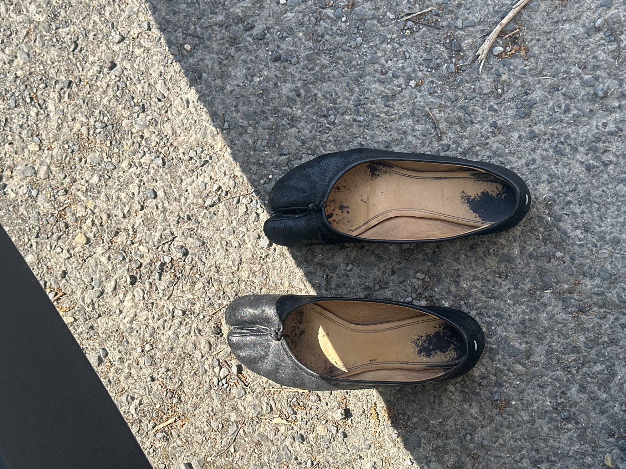 An overhead shot captures two black tabi-style ballet flats resting on a rough, grey paved surface, likely asphalt or concrete, outdoors in Waverley, United Kingdom. The shoes are positioned diagonally across the frame, with the top shoe slightly higher than the bottom one. Both shoes feature a distinct split-toe design and appear well-worn, displaying scuffs and dirt, particularly on their light brown/tan inner soles.
Bright, direct sunlight illuminates the scene, casting sharp shadows that indicate a clear, sunny day, likely mid-day. A prominent dark, diagonal shadow stretches across the top left portion of the image, possibly from an unseen structure or object. Small stones, gravel, and sparse dry debris are scattered across the ground, hinting at an untidy or natural outdoor setting. No people or activities are visible, and there is no discernible text within the image. The overall impression is one of discarded or temporarily placed footwear in a mundane outdoor environment.