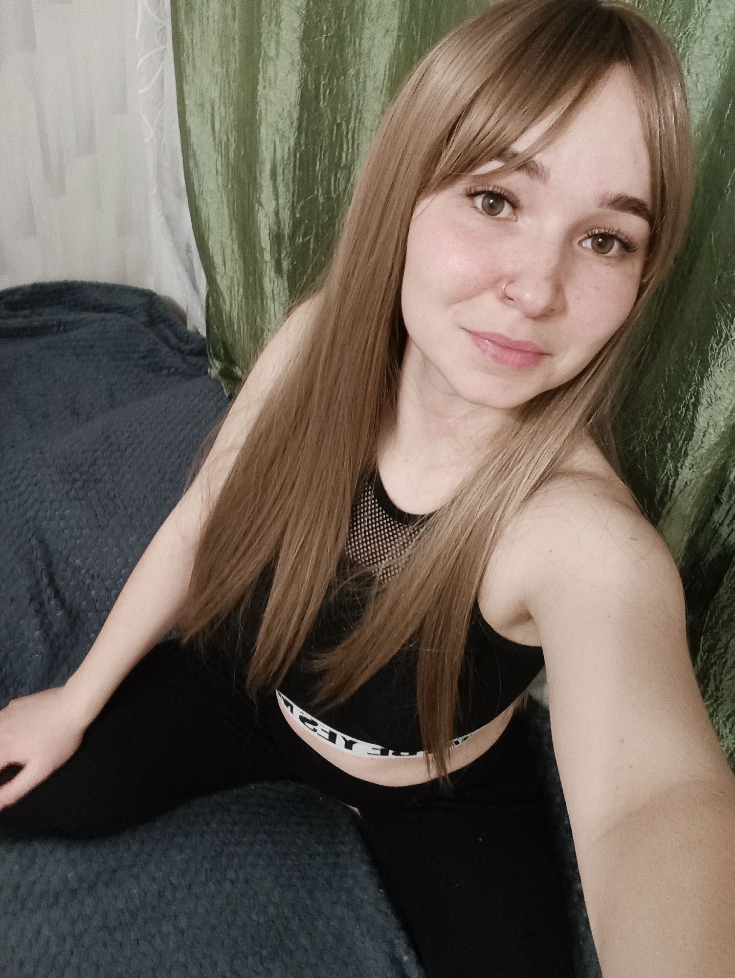 A female individual is depicted in a close-up, self-portrait style indoor setting. She has light-toned skin, light brown hair with bangs, brown eyes, and several freckles across her nose and cheeks. A small silver nose ring is visible on her left nostril, and her lips are painted with light pink lipstick. She is wearing a black athletic crop top featuring a mesh panel over the upper chest and collarbone area. Below the crop top, the white waistband of black leggings or pants is visible, displaying reversed text that appears to read "YES". The individual is seated, looking directly at the camera, with her left hand resting on the surface next to her. The immediate environment consists of a dark blue/grey textured fabric surface, likely a blanket or bedspread, on which she is sitting. The background features a light-colored, possibly white, textured wall or curtain on the left, adjacent to a dark green, reflective, and wrinkled curtain on the right. The specific location within the city of Izhevsk, Russia, cannot be determined from the visual information provided in the image.