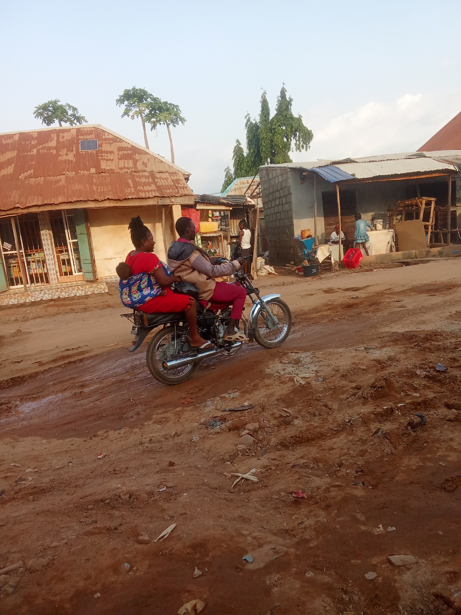 Two individuals, a man and a woman, are riding on a motorcycle on a dirt road in Kagini, Nigeria. The woman is seated behind the man, and a young child is strapped to her back with a colorful cloth. The motorcycle is traveling from left to right across the frame. The environment appears to be a residential or commercial area with buildings constructed from corrugated metal and cinder blocks. One building on the left has a rusted, corrugated metal roof and barred windows. Several trees are visible in the background. The road is unpaved and appears to be muddy in places, indicating recent rainfall. The sky is clear with a few scattered clouds. The scene is captured in the late afternoon or early evening light, as suggested by the warm, soft illumination.
