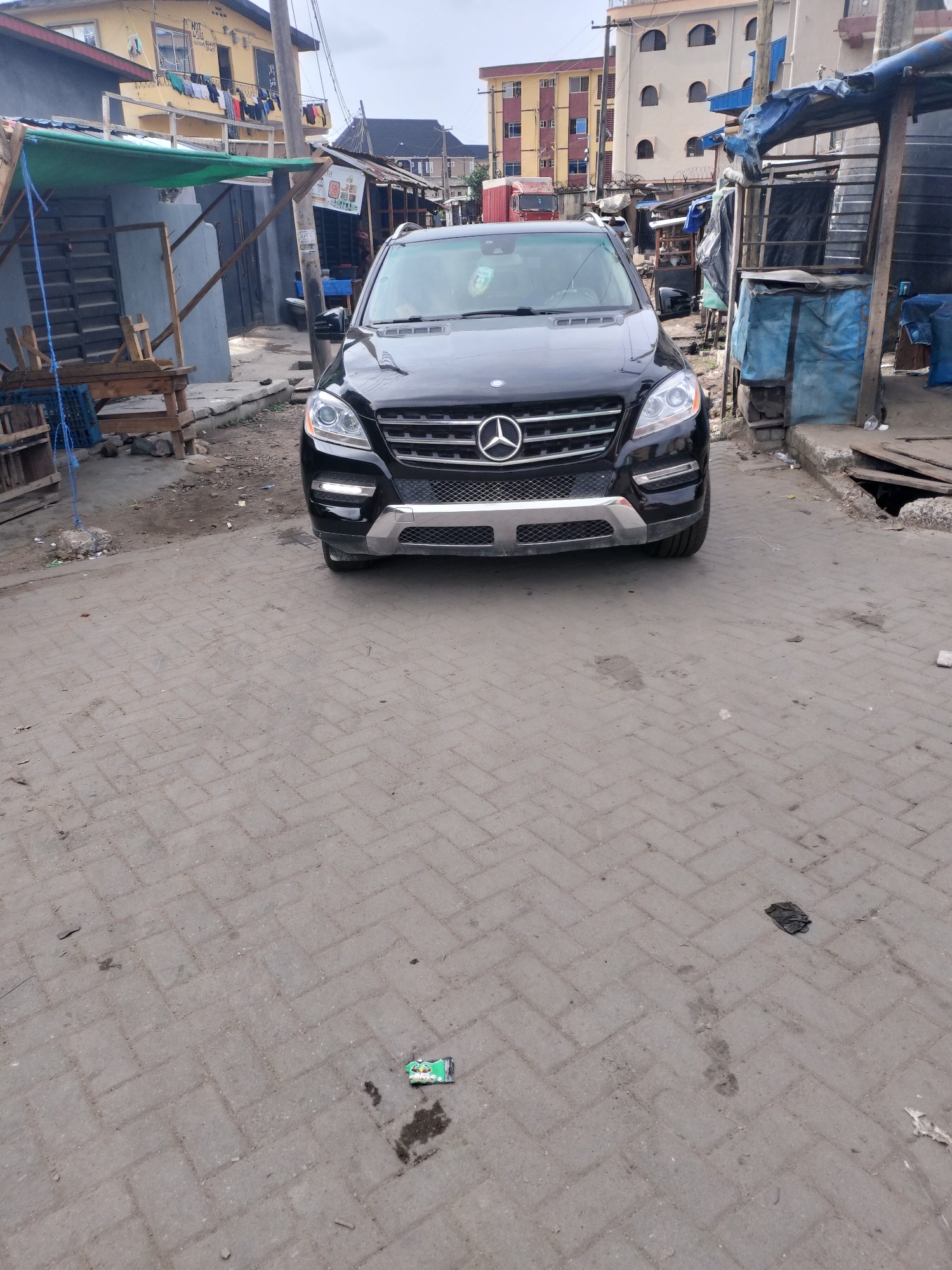This media file depicts a static urban street scene in Alaiyabiagba, Nigeria, under an overcast sky during daytime.
A black Mercedes-Benz ML-Class SUV is prominently parked in the center foreground, facing the viewer, on a street paved with grey interlocking bricks. The pavement shows signs of wear, dirt, and scattered debris, including several dark stains and a flattened green wrapper in the immediate foreground.
In the background, on the left side, stands a two-story building with a faded yellow facade. Laundry is visibly hanging from lines on its balconies. Below this building, a makeshift commercial stall or structure, constructed from wood and covered with a green tarp, is visible. Blue ropes are tied around its wooden supports, and some wooden crates are near its base. Faintly visible text on a white sign on the yellow building reads "NUT ASIDE."
On the right side of the image, another multi-story building with arched windows can be seen further back. Closer to the foreground, a second makeshift structure, possibly another stall or storage area, is covered with blue tarpaulins. Large black barrels, likely water tanks, are positioned behind this structure. An open drainage ditch or hole, partially covered by wooden planks, is visible on the far right edge of the paved street. The scene portrays a busy, lived-in environment characteristic of an urban area with mixed residential and commercial activity.