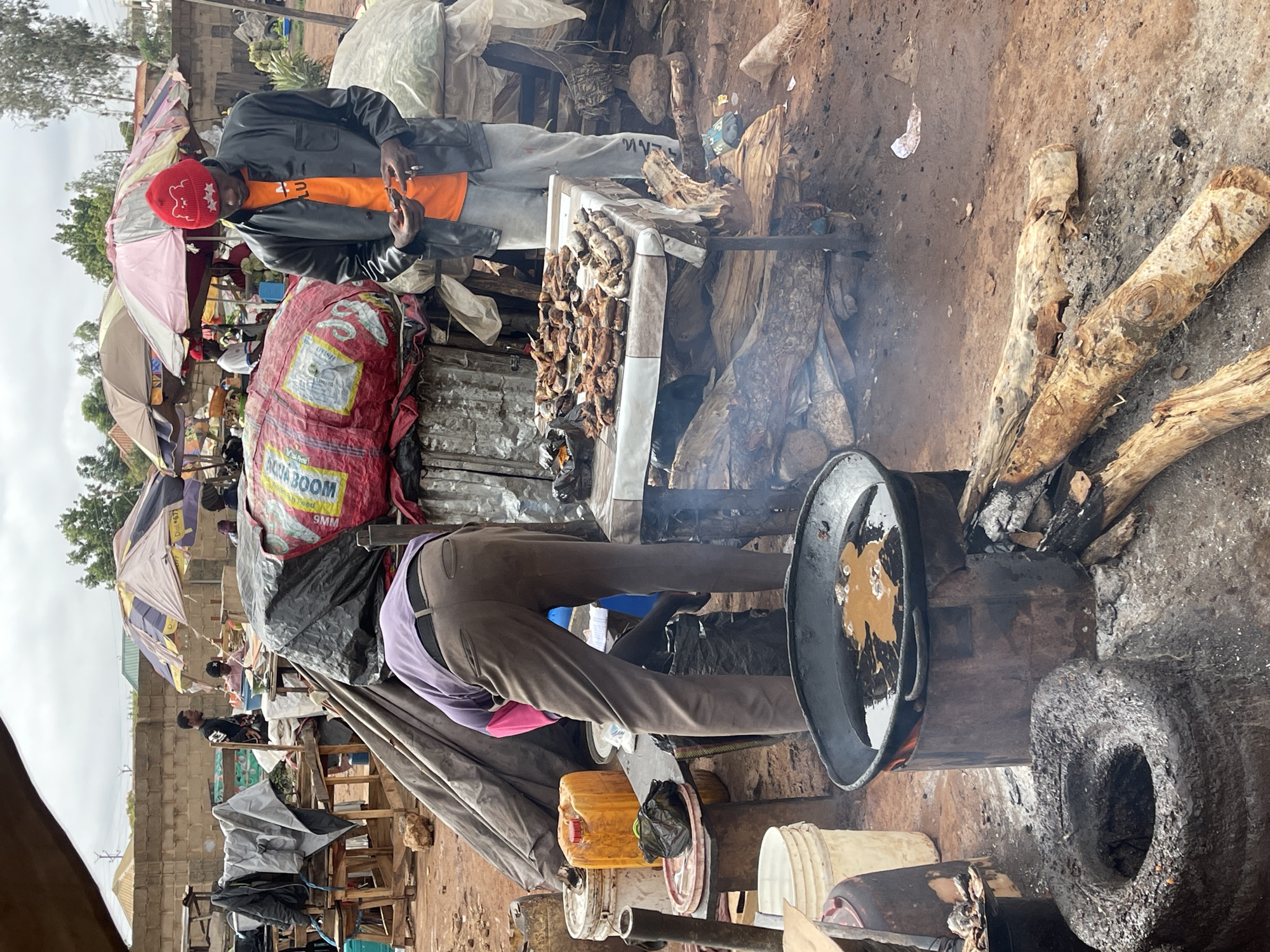 This image depicts an outdoor food vending operation in Gura Top, Nigeria. A man in a black leather jacket and red hat is standing and talking on his phone near a table laden with grilled meats. Another individual is bent over, appearing to tend to a large frying pan or cooking pot heated over a wood fire built on makeshift supports, including what appears to be an old tire. The scene is cluttered with bags, containers, and cooking paraphernalia. Other people are visible in the background amongst the makeshift market stalls and umbrellas. The environment appears to be a dusty, informal marketplace.