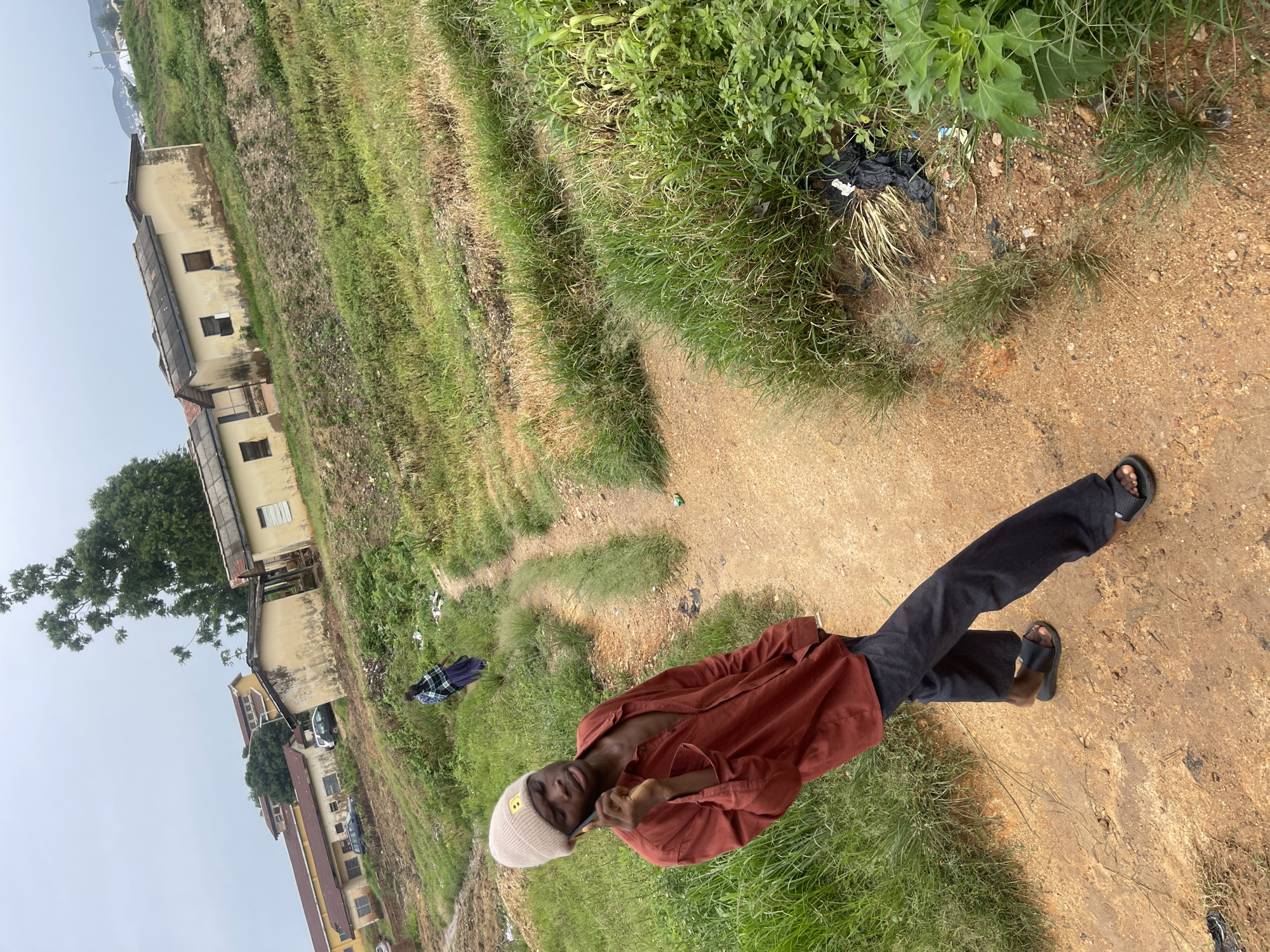 A young man wearing a maroon unbuttoned shirt, dark pants, a beige beanie, and black slide sandals is standing on a dirt path, holding a cell phone to his ear. He is positioned in the foreground, facing the camera at a slight angle. Behind him, another person in a plaid shirt and dark skirt walks away up the path. The path winds through a grassy, uneven terrain with some scattered debris. In the background, several weathered, light-colored buildings with corrugated metal roofs are visible on a hillside. The sky is overcast. The scene appears to be in an outdoor, semi-rural or underdeveloped urban setting, possibly a residential area on the outskirts of a city.