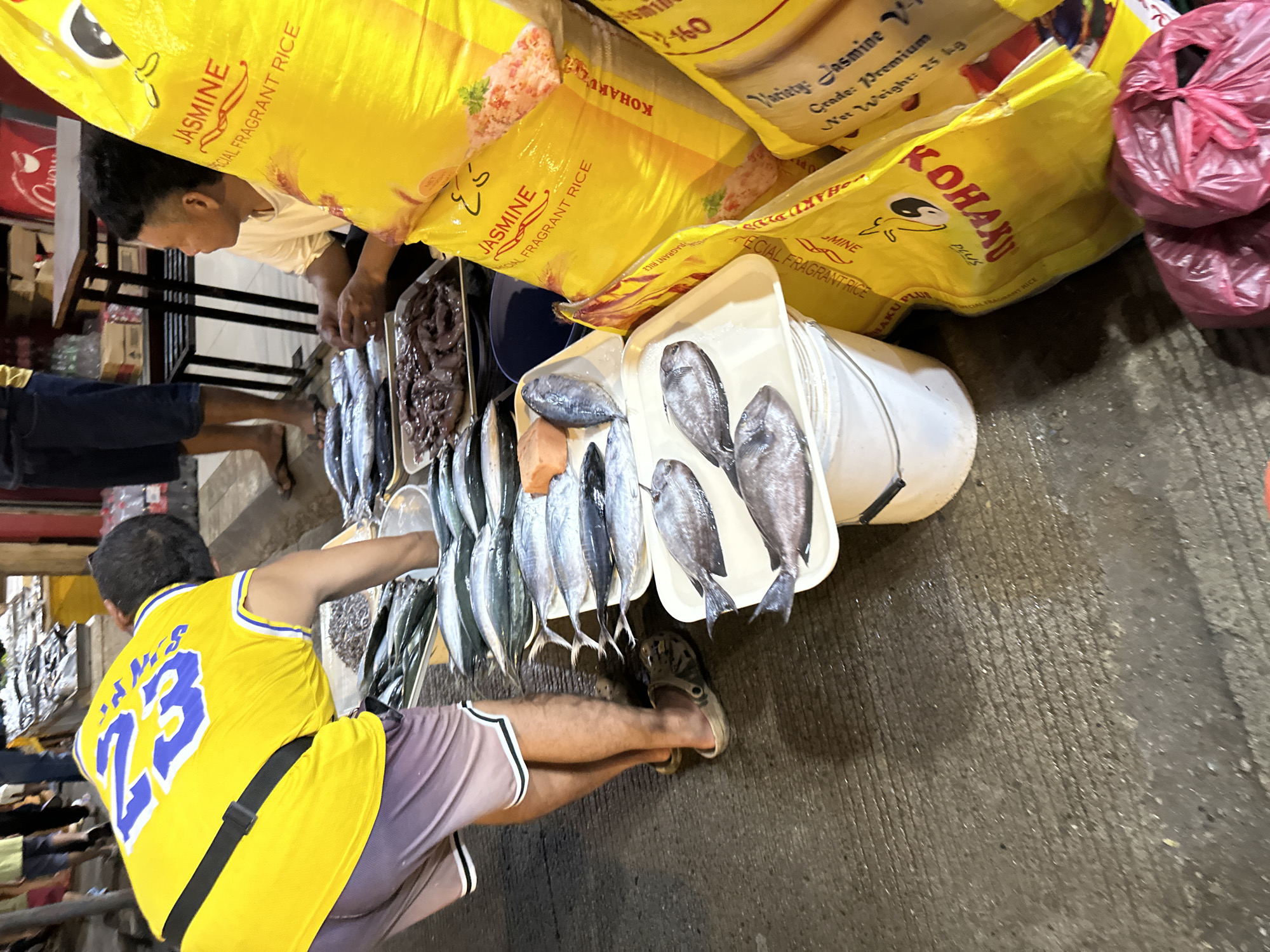 In a wet market in Davao City, Philippines, a man wearing a yellow jersey with the number 23 and purple shorts is bent over a display of fresh fish. He is positioned on the left side of the frame, with his back to the camera, reaching towards the fish. Several trays of assorted fish, some whole and some filleted, are laid out on the ground and on top of white buckets. In the background, another man in a white t-shirt is seated, looking towards the right. Large yellow bags of "JASMINE SPECIAL FRAGRANT RICE" are prominently displayed to the right of the fish, some open and partially visible. The ground appears to be concrete and wet, indicating a typical market environment. The overall scene depicts a commercial transaction or preparation of seafood for sale.