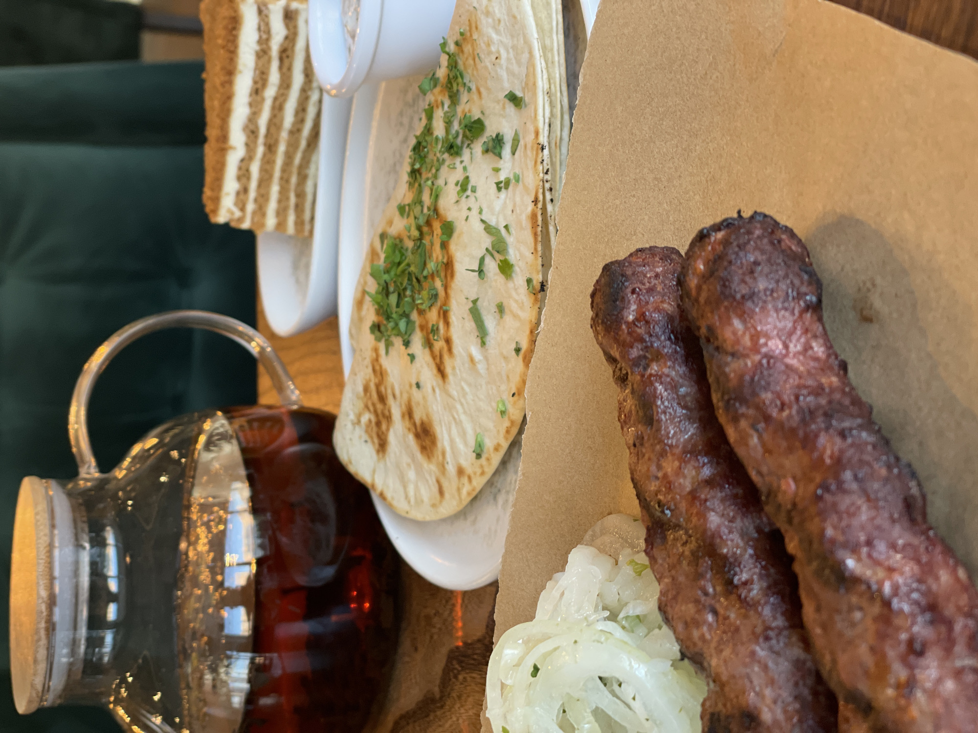 A close-up, eye-level shot captures a meal at a restaurant in Kazan, Russia. In the foreground, two grilled meat skewers, likely lamb or beef, are arranged on a piece of brown paper. To their left is a pile of thinly sliced white onions, perhaps pickled or raw. Behind these, a flatbread, lightly browned and topped with chopped green herbs, rests on a white plate. A small white ramekin containing a creamy sauce sits to the right of the flatbread. In the background, a glass teapot filled with dark red liquid, possibly tea, is positioned on the left. A slice of layered cake, with visible cream and brown cake layers, is presented on another white plate to the right of the teapot. The setting appears to be a restaurant with dark green upholstered seating visible in the background, suggesting a cozy and somewhat formal atmosphere. The lighting is warm, highlighting the textures and colors of the food.