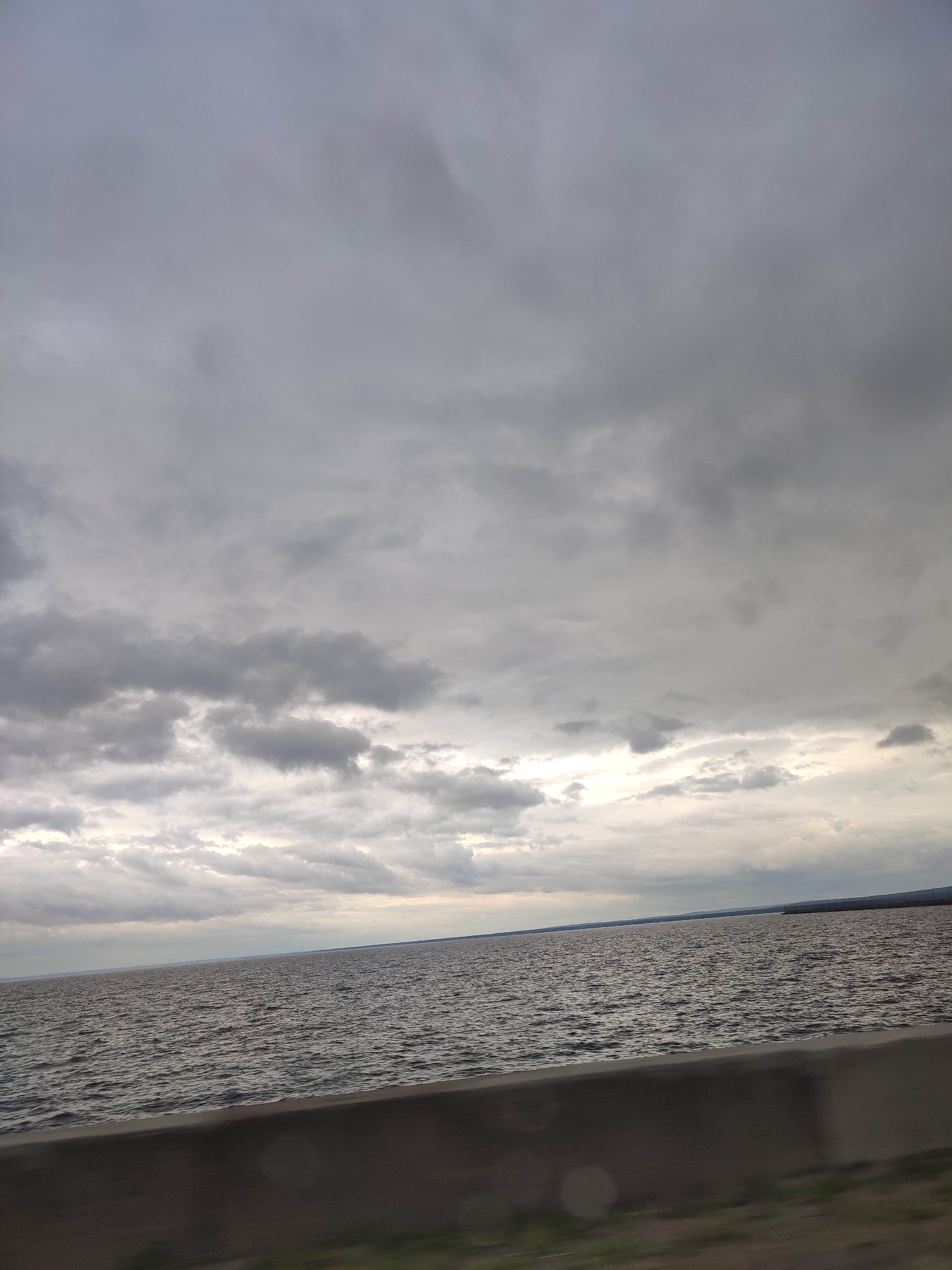 The image displays an expansive, dark body of water, likely a large lake or sea, under a heavily overcast sky. The upper approximately two-thirds of the frame are dominated by dense, mottled gray clouds, with streaks of brighter, diffused light visible along the lower portion of the sky near the horizon line. The water surface is dark and exhibits numerous small, irregular ripples, suggesting wind activity. A faint, dark landmass is discernible on the distant horizon towards the right side of the frame. In the immediate foreground, a dark gray concrete barrier or wall runs horizontally across the bottom approximately 15% of the image, appearing to be a roadside divider or embankment. Below the barrier, a blurry strip of dark ground with hints of green vegetation is visible. The scene is devoid of identifiable people, animals, or distinct man-made structures beyond the foreground barrier. The lighting is uniform and muted, consistent with a cloudy day. The specific location in the city of Unknown, Unknown, is unidentified.