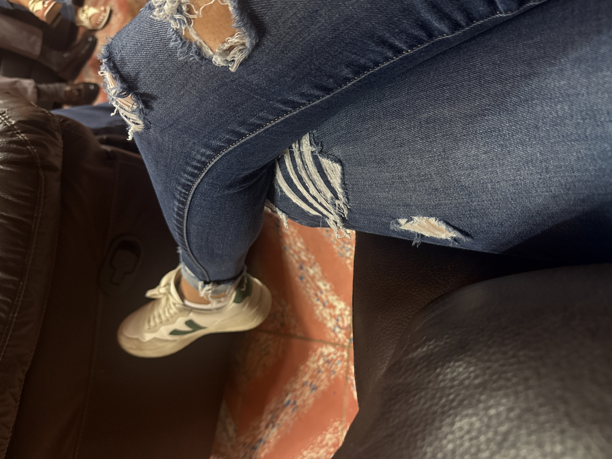 A close-up, top-down shot depicts a person seated, wearing distressed blue jeans with multiple rips and tears exposing skin. The person is wearing a white sneaker with a green logo on the side. The individual is seated on an armchair, possibly a sofa, with brown leather upholstery visible on either side of the image. The floor is decorated with a patterned, red-toned tile.
The image location is San Cristobal, Mexico.