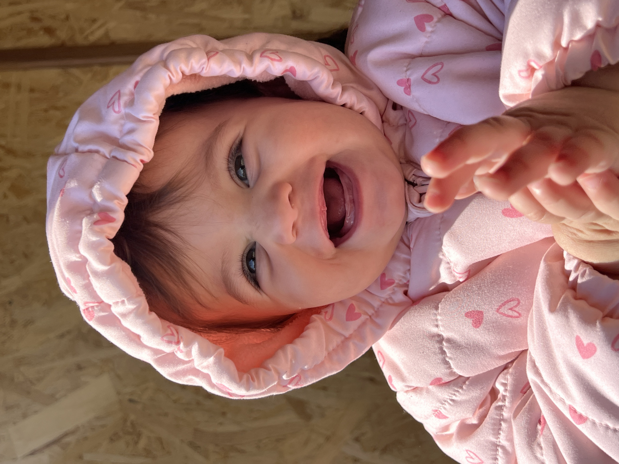 The image is a close-up shot of a baby lying on its back. The baby is wearing a pink hooded jacket with heart patterns and is smiling with its mouth open. The baby's eyes are visible, and it appears to be reaching out with its right hand. The background appears to be a wooden surface. No information is available about the location being in the city of Elena, Bulgaria.