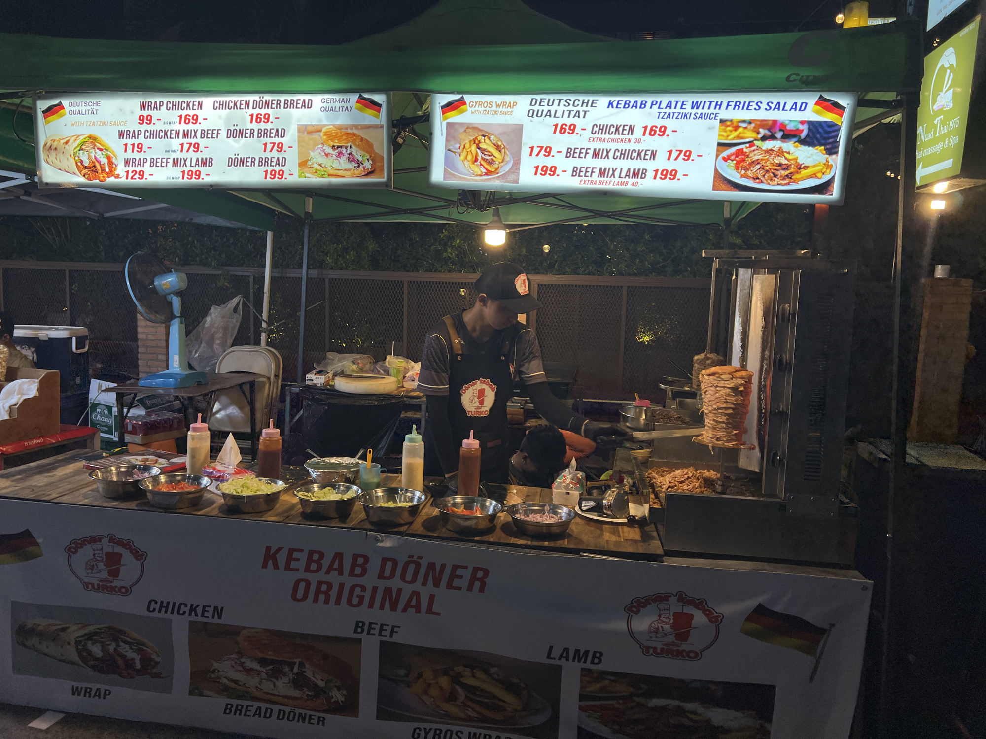 This image shows a Kebab Döner Original food stall in Baan Bo Phut, Thailand. The stall is set up under a green canopy, with bright menu boards displaying German-style kebab wraps, döner bread, gyros wraps, and kebab plates with fries and tzatziki sauce. Prices are listed in Thai Baht.
Behind the counter, a man wearing a cap and apron is working at the stall. He appears to be preparing or serving food. Various ingredients such as chopped onions, tomatoes, lettuce, and sauces are set out in bowls on the counter. To the right, a vertical rotisserie is visible with döner meat cooking.
The front of the stall is decorated with images of the food items and logos of "Döner Kebab Turko", along with German flags. In the background, there is a fence, some foliage, and another sign for "Nuad Thai 1976 Thai Massage & Spa".