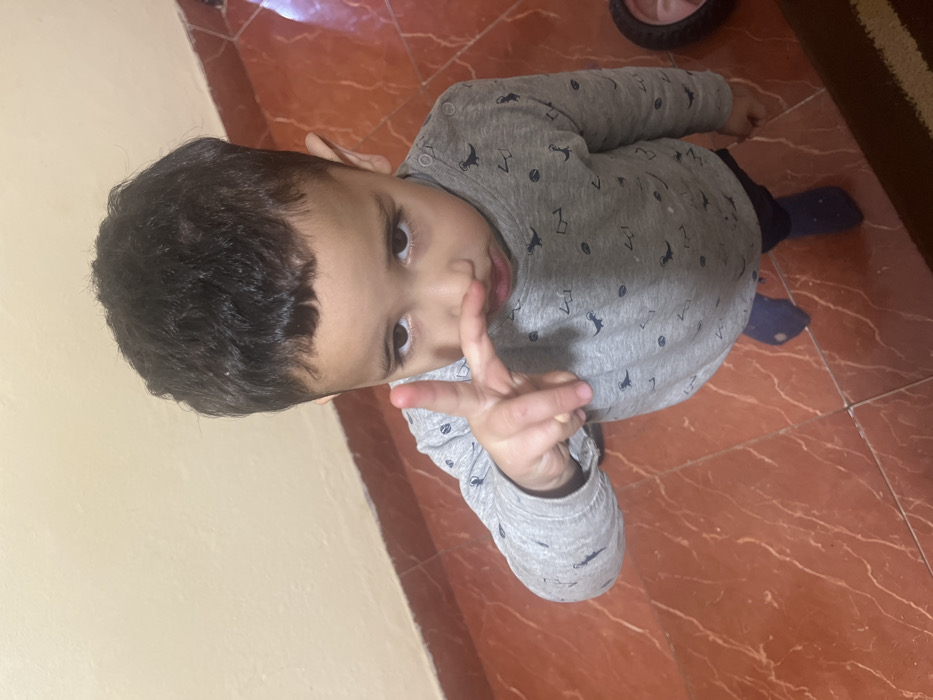 This image depicts a young male child indoors, located in Ouaouizert, Morocco. The child, with dark, short, curly hair and dark eyes, is positioned facing the camera from a slightly elevated angle. He is wearing a long-sleeved grey shirt patterned with dark dinosaur figures and other shapes, dark blue pants, and blue socks. His right hand is raised, with the index and middle fingers extended forward and slightly bent, and the thumb extended below them, forming a distinct hand gesture. The background consists of a plain light beige wall above a dark reddish-brown baseboard. The floor is covered with glossy, reddish-brown tiles featuring irregular lighter vein patterns, with visible light-colored grout lines. A portion of a pink wheel, possibly from a toy, is visible on the far right side of the frame.