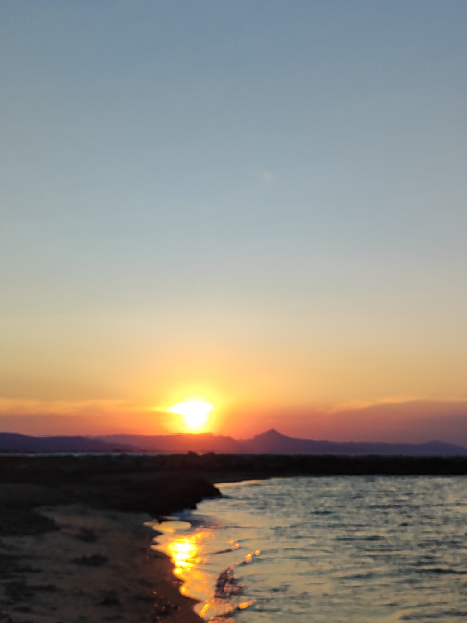 The image depicts a serene coastal scene at sunset in Dénia, Spain. The sun, a brilliant orb of light, is setting low on the horizon, casting a warm, golden glow across the sky and water. The sky transitions from a pale blue at the top to hues of orange and pink as it nears the horizon, with some soft clouds visible.
In the foreground, a sandy beach meets a body of calm water. The gentle waves reflecting the sunlight create shimmering paths of light on the water's surface. The shoreline is dark and indistinct, suggesting the presence of vegetation or rocks. In the distance, a silhouetted mountain range forms a rugged backdrop against the colorful sky.
The overall mood of the image is peaceful and tranquil, characteristic of a sunset by the sea. The absence of people or man-made objects emphasizes the natural beauty of the location. The time of day is clearly sunset, indicated by the sun's position and the warm colors in the sky. The location cues, such as the coastal landscape and the distant mountains, are consistent with the Spanish coast. There is no visible text in the image.