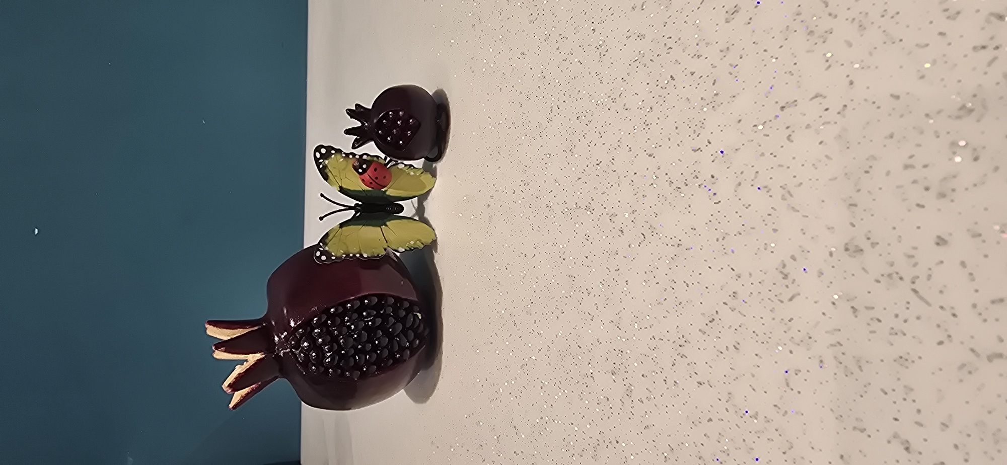 The image shows a decorative item featuring two pomegranates and a butterfly with a ladybug on its wings. The pomegranates are a deep red or burgundy color, with exposed dark seeds in one of them. The butterfly has bright green wings with black dots and a red body with black spots, resembling a ladybug. The decorative item appears to be attached to a wall, which is partially visible on the left side as a solid teal color. The right side of the wall has a white surface covered in a fine glitter or sparkly texture. The lighting suggests it is indoors. There is no text visible in the image. The scene is static and focuses on the decorative elements.