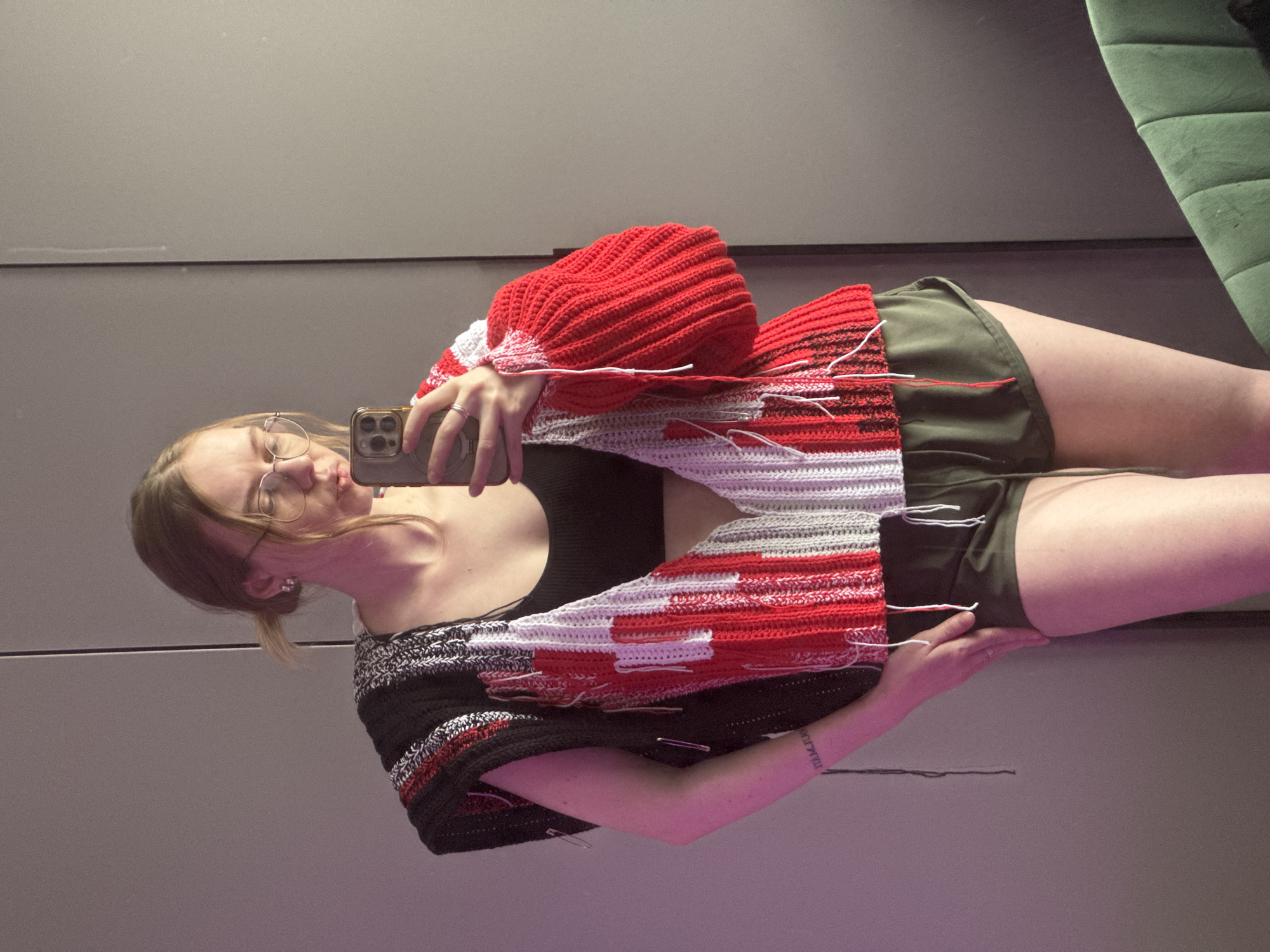 A young woman is standing in what appears to be an interior space, possibly a dressing room or a large closet, taking a selfie in the mirror. The background is a wall with horizontal panels. The woman is wearing a black tank top and green shorts. She is holding her cell phone up to take the picture. She has on a pair of glasses. She is wearing a red, white, gray, and black crocheted shawl/sweater with some loose yarn ends visible. There is a glimpse of a green cushion or part of furniture in the upper right corner of the frame. The photo appears to be taken in a mirror, as the image is reversed. The woman has a tattoo on her left wrist. There is no information available to confirm the image was taken in Krasnoyarsk, Russia.