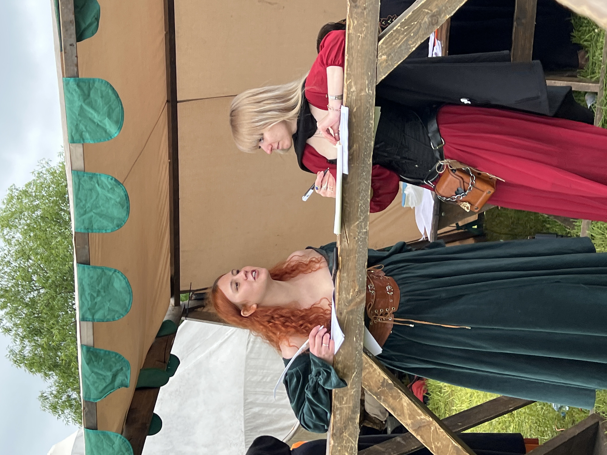 Two women are standing beneath a tent structure with a beige canopy and green trim. One woman, with red curly hair, is wearing a long green dress and holding papers. The other woman, with blonde hair, is wearing a red dress with a black corset-style top and is writing on a paper with a pen. Both women are dressed in what appears to be historical or fantasy-themed attire. The tent structure is made of rough-hewn wooden posts. The background appears to be a grassy area, and the sky is overcast. The location is Unknown, Russia.