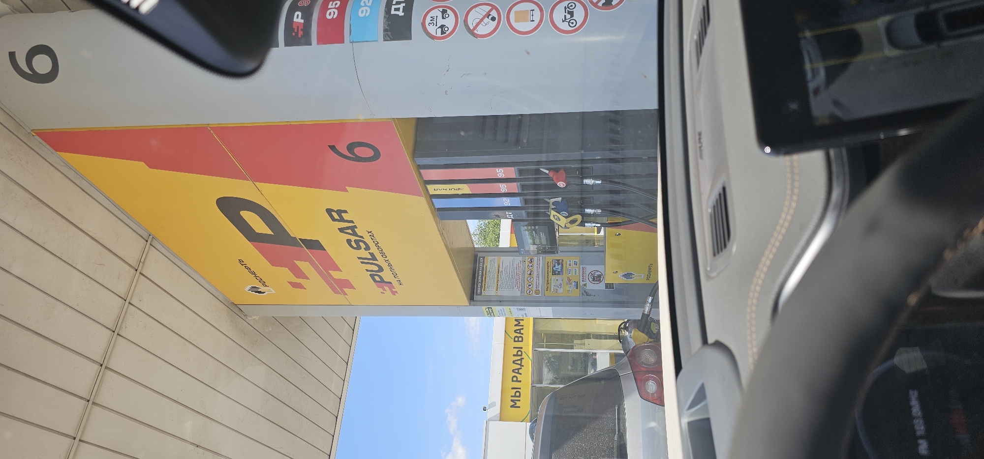The image shows a close-up of a gas station. The sign for "HP PULSAR" is visible, along with the number "6" on a column. There are gas pumps with different colored nozzles, and a sign with prices including "92" and "95". A car is parked at one of the pumps, with its rear visible. In the background, a yellow building with the text "МЫ РАДЫ ВАМ" (We are glad to see you) is visible. The overall environment appears to be an outdoor gas station. The image was taken from inside a vehicle, as indicated by the dashboard and windshield. The location is identified as Ovoshchnoy, Russia.