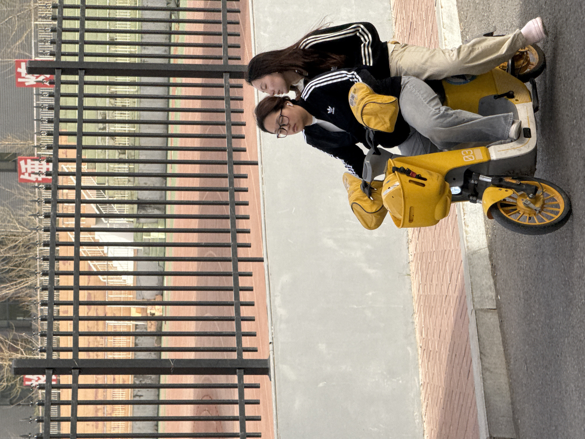 Two young women are riding on a yellow scooter on a paved surface. The woman driving wears glasses, a white t-shirt, a black Adidas track jacket with white stripes on the sleeves, and gray pants. Her passenger is also wearing a black Adidas track jacket with white stripes on the sleeves and beige pants. The scooter has yellow side bags. Behind them is a fence that appears to be overlooking a sports field. Red signage can be seen through the fence. The setting appears to be an urban environment, potentially Zhongtun, China.