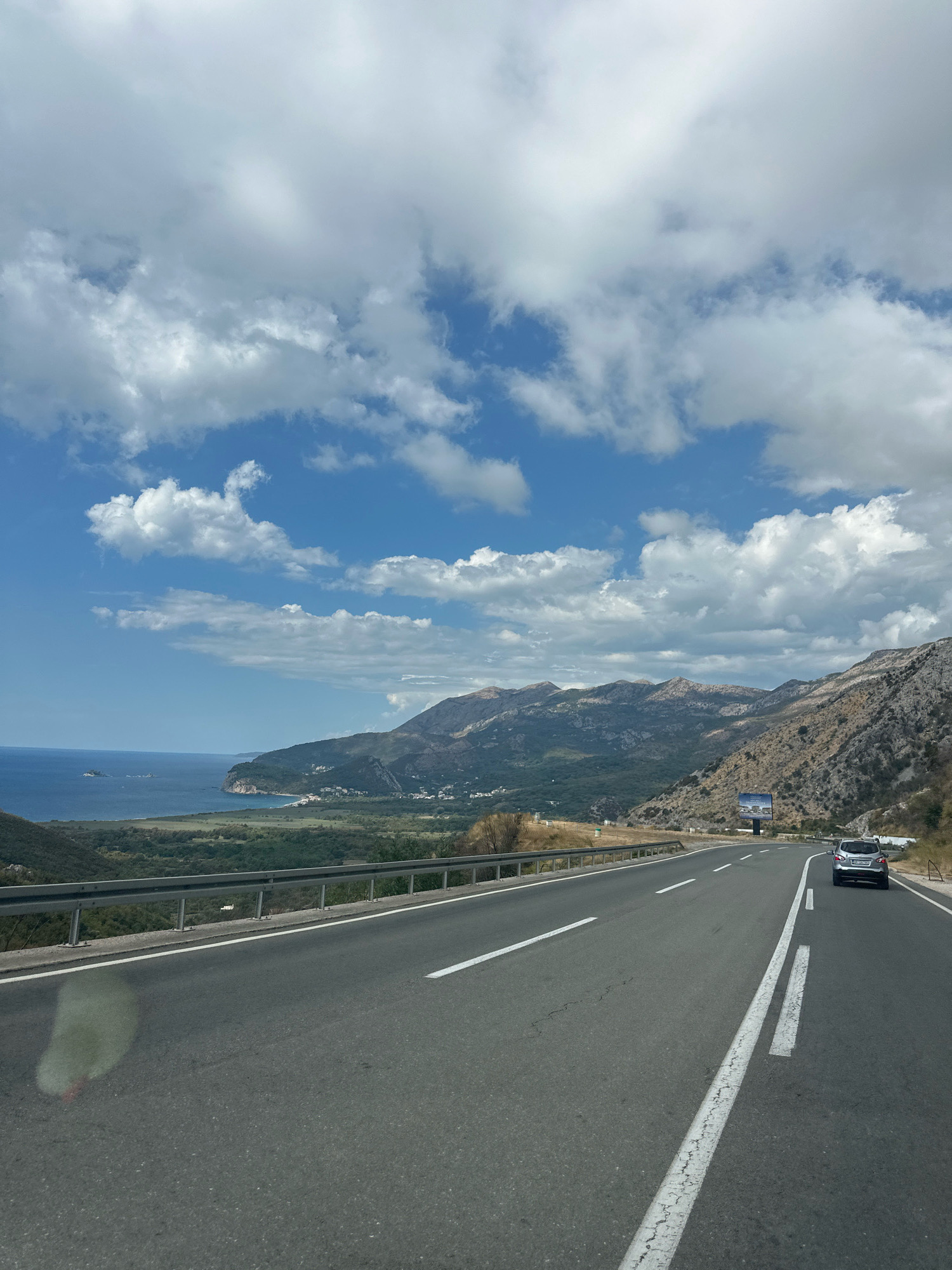 A scenic view of a coastal highway in Kanjoši, Montenegro, captures a winding road bordered by a metal guardrail. On the right side of the road, a silver car drives ahead, its license plate visible. Beyond the road, a steep, rocky mountain range dominates the landscape, with sparse vegetation clinging to its slopes. In the distance, the azure sea meets a rugged coastline dotted with small settlements. A lush green valley separates the mountains from the coast, further enhancing the picturesque scene. The sky above is a vibrant blue, adorned with fluffy white clouds, suggesting fair weather. A billboard stands on the side of the road, adding a touch of man-made structure to the natural beauty.