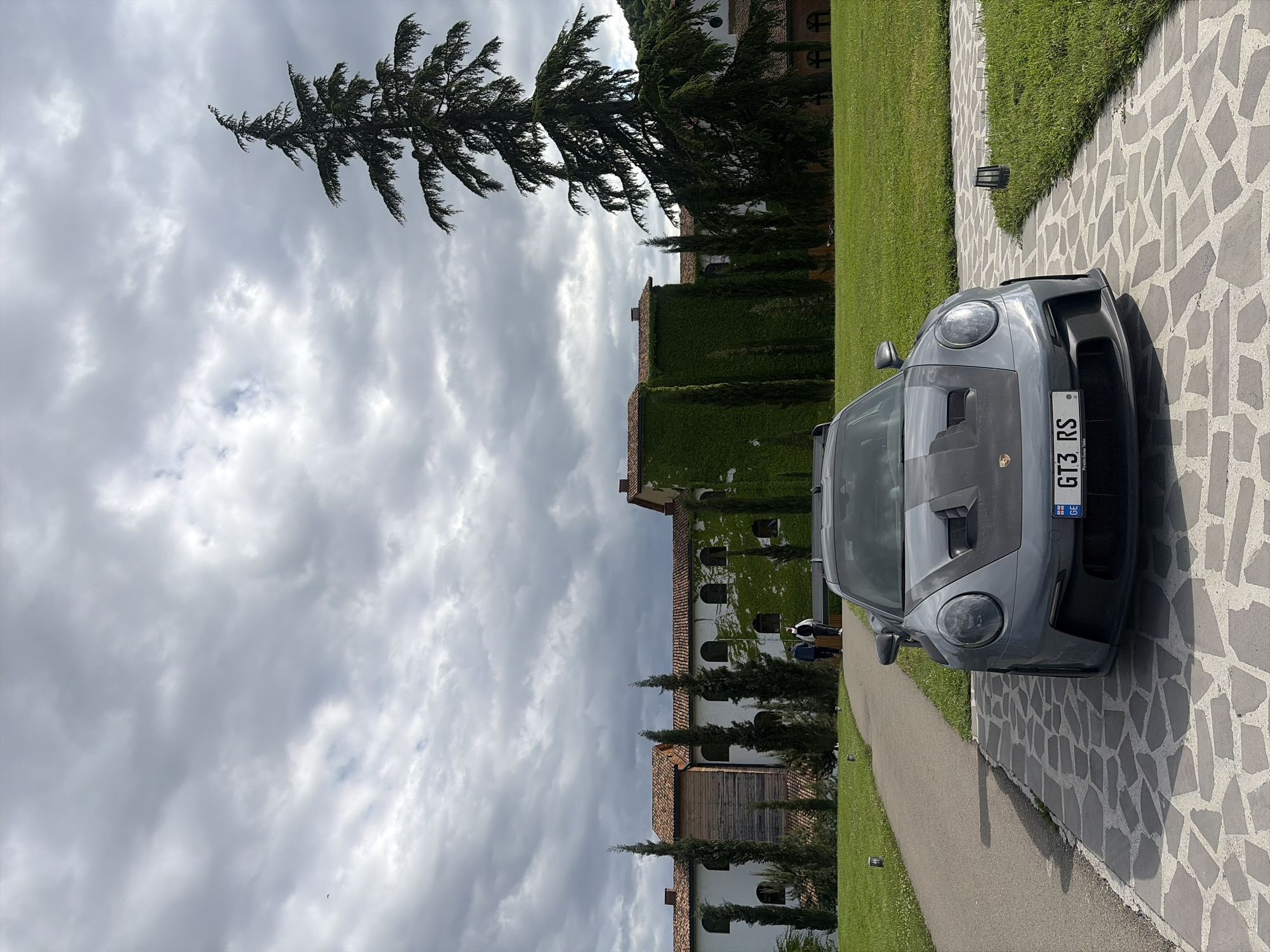 The image captures a grey Porsche 911 GT3 RS parked on a patterned stone pathway outside a stately building, under a cloudy sky.
In the foreground, the front of the Porsche 911 GT3 RS is visible, positioned slightly to the right, facing away from the immediate viewer. Its license plate clearly displays "GT3 RS" and "GE," indicating Georgia, with "Porsche Center Tbilisi" subtly visible below. The car's sleek, performance-oriented design, including its headlights and front air intakes, is prominent. The car is situated on an intricate pathway made of irregularly shaped grey and off-white stones. To its left, a meticulously maintained green lawn stretches towards a narrower dirt path.
The midground features a grand building with a terracotta-tiled roof. A large section of its facade is adorned with dense green ivy, giving it a classic and established appearance. Several tall, slender, dark green coniferous trees, resembling cypresses, line the property, adding to the picturesque landscape. A few indistinct figures of people are visible in the distance near the building.
The sky is largely covered with an array of grey clouds, suggesting an overcast day. The overall scene depicts a serene and upscale setting, likely an estate or resort, consistent with its location in Napareuli, Georgia. The time of day appears to be daytime, with ample natural light despite the cloud cover.