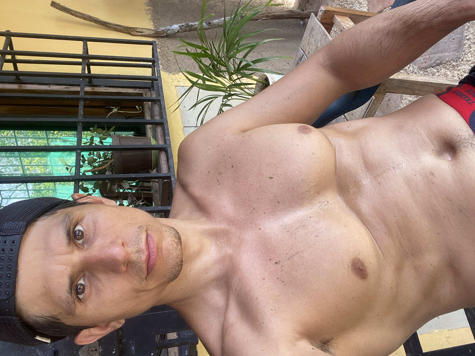 A shirtless man wearing a black cap is posing for a picture. The man has brown eyes, a light beard, and appears to be in an outdoor setting. A plant in a pot is visible in the background, as is a metal structure resembling a window grill. The background also suggests an outdoor environment. There is no evidence in the image to indicate the specific location of Los Velásquez, Venezuela.