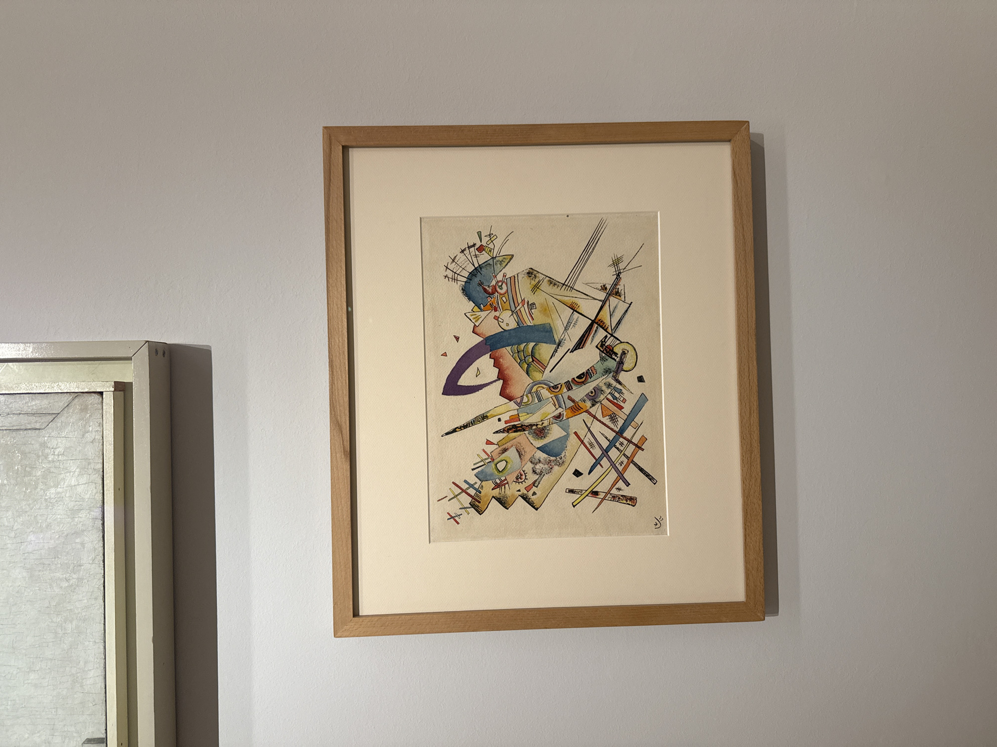 A framed abstract painting hangs on a gray wall next to a section of another painting. The abstract painting is framed in light brown wood, with a light beige matte and an irregularly shaped paper with the image. The abstract art consists of layered shapes, and lines in red, black, blue, yellow and other colors. Based on the style of the painting, it is possibly a work by Wassily Kandinsky. The location is presumed to be in Madrid, Spain. There are no people visible.