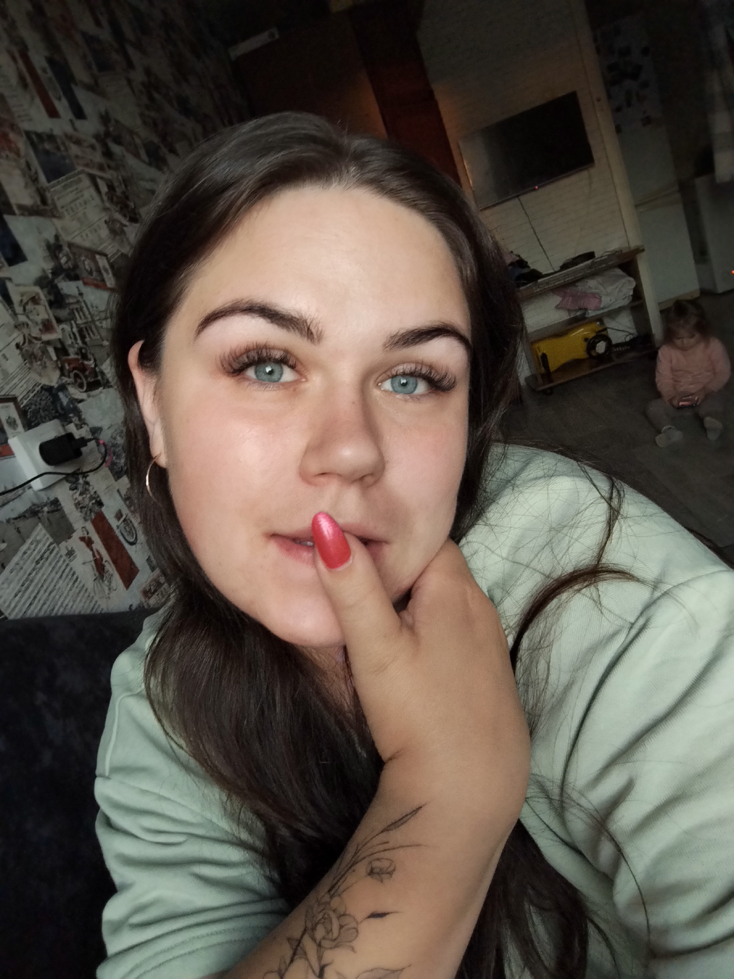 This is a close-up, eye-level selfie taken by a young adult woman indoors, likely within a home in Chaikovsky, Russia. The woman, with long dark hair, striking blue eyes, and prominent eyelashes, is the primary subject. She has well-defined eyebrows and wears a light green long-sleeved top. Her right hand, adorned with bright reddish-pink nail polish, is gently touching her lips. A tattoo with a floral or botanical design is visible on her right forearm. Her expression is calm and directs a gaze towards the camera.
In the background, to the left, a wall is covered with distinct wallpaper featuring a collage of vintage-style prints, possibly old advertisements or postcards with text and illustrations. A white electrical outlet is visible on this wall. To the right, further back, a wall-mounted flat-screen television is positioned above a wooden shelf unit holding various items, including a bright yellow object. The floor is dark.
A small child, possibly a toddler, is seated on the dark floor in the distant background, wearing a light pink top and grey pants. The child appears to be quietly engaged with a small object, possibly a phone or toy. The lighting in the room is dim, suggesting an evening or nighttime setting. No specific text is clearly legible on the wallpaper or other objects.