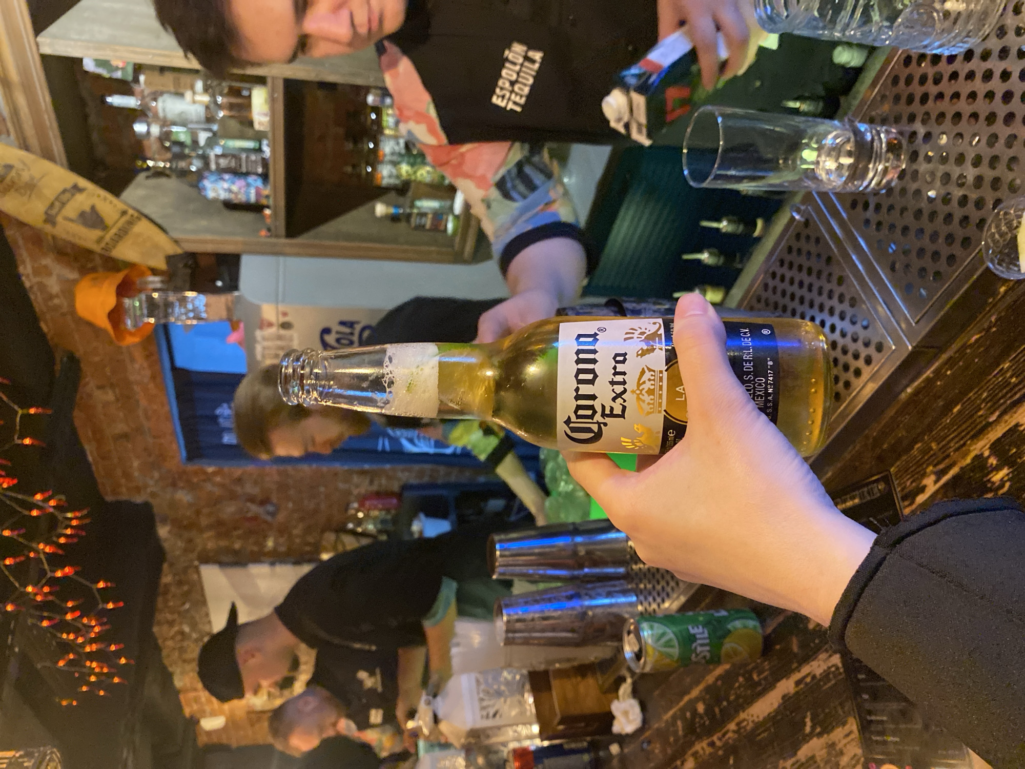 A person is holding a bottle of Corona Extra beer. The beer is foamy at the top. The label is visible with the text "Corona Extra". A bartender is in the background, wearing a black apron that has the text "ESPOLON TEQUILA". In the background, there is a well-stocked bar with bottles of liquor. Clear drinking glasses are on the bar. The location appears to be a bar, possibly in Izhevsk, Russia, although this can't be confirmed from the image alone.