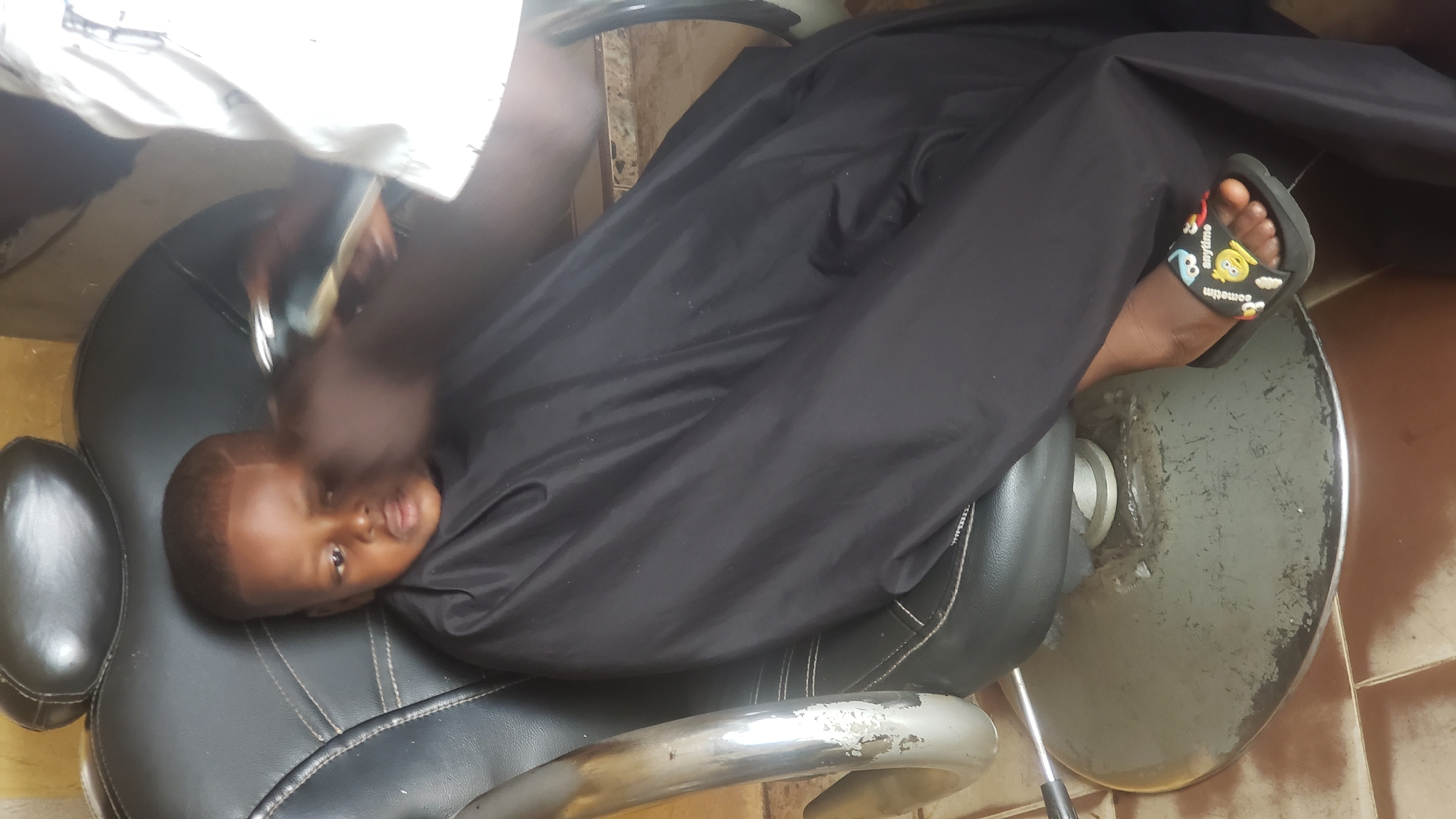 A young child, lying back in a black barber's chair with a black cape draped over them, receives a haircut. A barber is using clippers. The child is wearing cartoon-themed sandals. The chair is in a barber shop location in Narayi, Nigeria. The floor appears to be tiled.