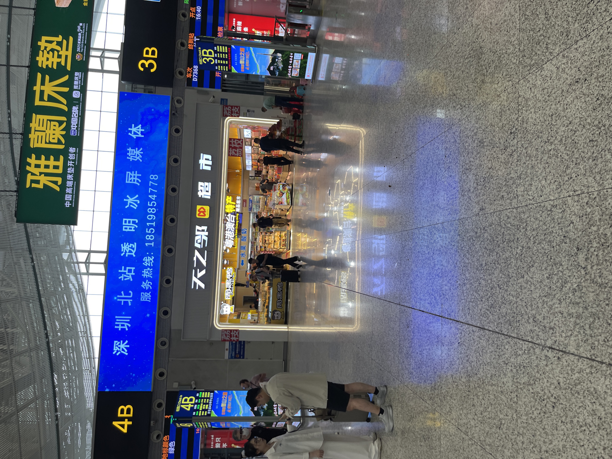 The image depicts the interior of a modern transportation hub, likely a train station or airport, in Longhua District, China. Large digital displays are prominent, with one blue screen showing "深圳·北站透明冰屏媒体" and "服务热线: 18519854778", indicating Shenzhen North Station and a hotline number. Another large green sign displays Chinese characters, likely an advertisement. Yellow "4B" and "3B" labels mark different sections or platforms.
In the foreground, two individuals, a woman in white attire and a man in a light-colored jacket and shorts, are engaged in a conversation while standing near luggage. Other people are visible in the background, some walking with luggage, others interacting with staff or browsing at a retail establishment labeled "天天乐超市" (Tian Tian Le Supermarket). The floor is tiled with a speckled pattern, reflecting the overhead lighting and digital displays, creating colorful light patterns. The environment appears clean and well-lit, consistent with a public transit facility.