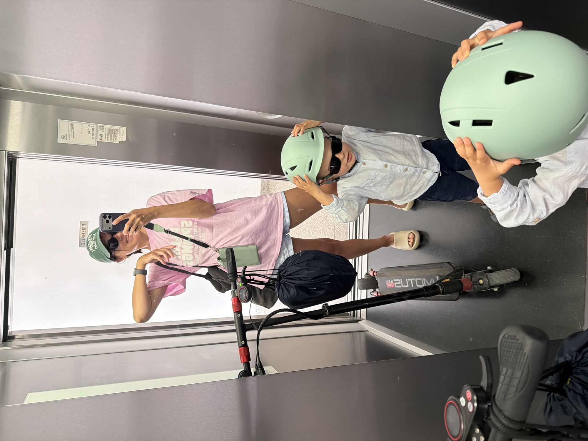 A woman wearing a pink t-shirt, denim shorts, a green baseball cap, and sunglasses is taking a selfie in a mirror. She is in an elevator with her child, who is wearing a light blue collared shirt, dark shorts, and sunglasses. They both are holding light green helmets. An electric scooter is also present in the elevator. The elevator's wall features informational stickers. The location is not identified, and I cannot determine if the image was taken in the city of Budva, Montenegro.