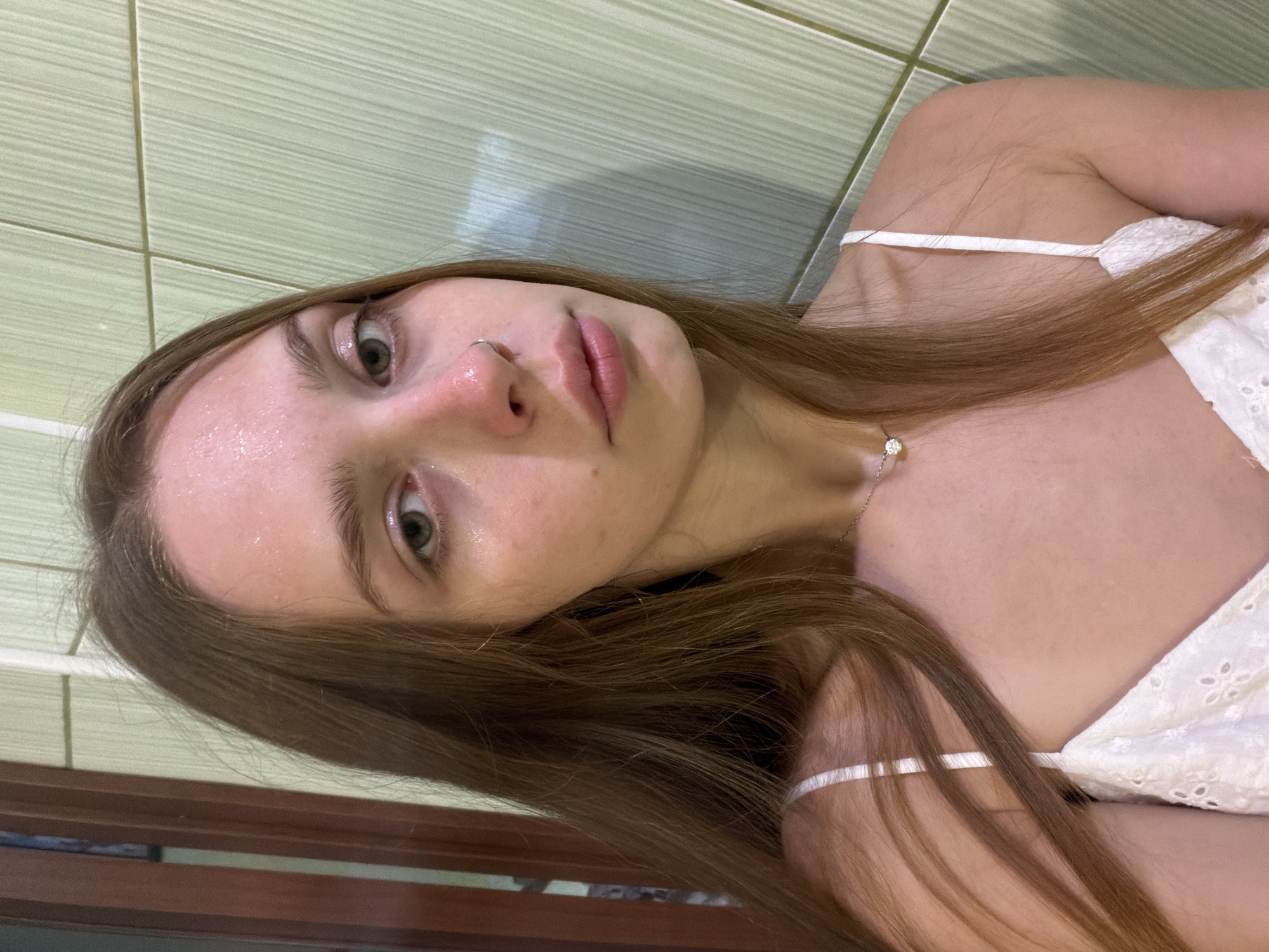 A young woman with long, brown hair lies on her side, her head tilted to the side. She wears a white, embroidered top with thin spaghetti straps. Her face is pale and slightly flushed, with a few droplets of sweat visible on her forehead and cheeks, suggesting warmth or exertion. Her eyes are a light green, and she has a small nose ring. Her lips are slightly parted. The background appears to be a tiled wall in a muted green color. The lighting is soft, casting subtle shadows. The overall impression is one of intimacy and a moment of rest or reflection. There is no visible text in the image.