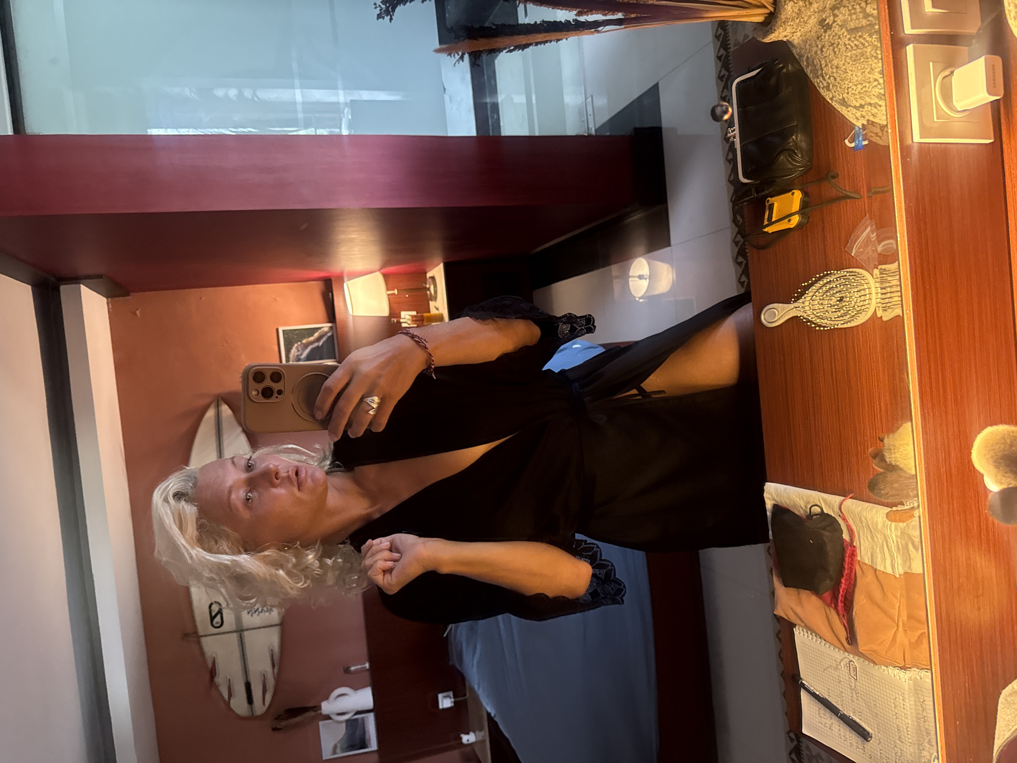 A woman with blonde, curly hair is reflected in a mirror, taking a selfie. She is wearing a black robe that is open, revealing her chest and part of her leg. She is also wearing a ring on her left hand. The room appears to be a bedroom, with a bed and a dresser visible in the background. There is a surfboard hanging on the wall behind the woman. The dresser in the foreground has several items on it, including a notebook, a brush, a purse, and what appears to be a makeup bag.