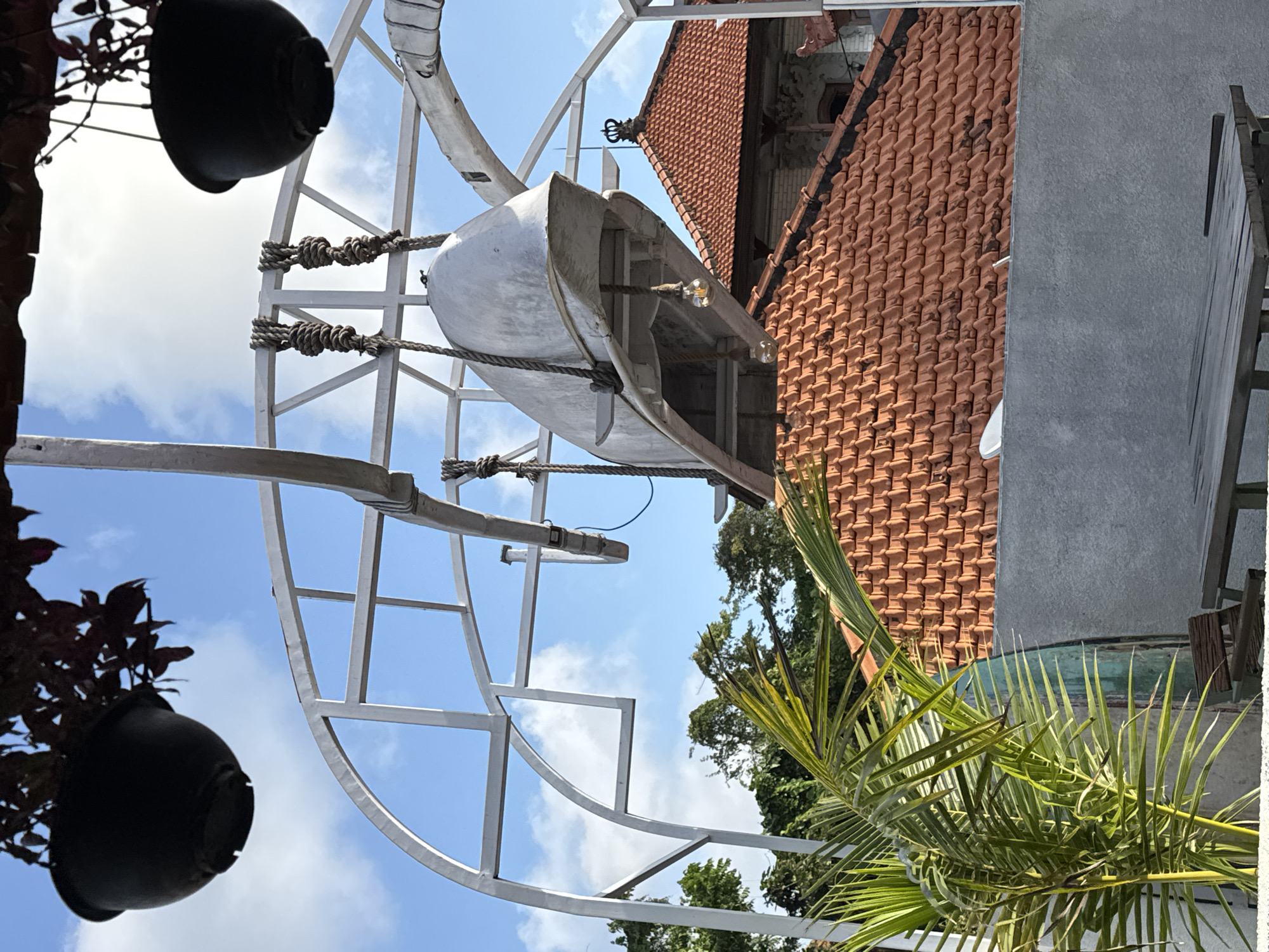 In Kuta Selatan, Indonesia, an outdoor scene unfolds under a blue sky with scattered clouds. A large, metallic structure, possibly an art installation or a decorative element, arches over the foreground. Suspended from this structure by thick ropes is a long, light-colored object that resembles the hull of a boat or a large fish sculpture. Two exposed light bulbs hang from the underside of this suspended object.
Below the metallic structure and the suspended object, a series of red-tiled rooftops are visible, characteristic of Indonesian architecture. A palm tree with vibrant green fronds is positioned in the lower left of the frame, partially obscuring a grey concrete wall. In the far background, lush green trees and foliage can be seen, suggesting a tropical environment. In the lower foreground, a simple wooden table and bench are set, hinting at an outdoor seating area. The overall impression is of a tranquil, perhaps artistic, outdoor space.
