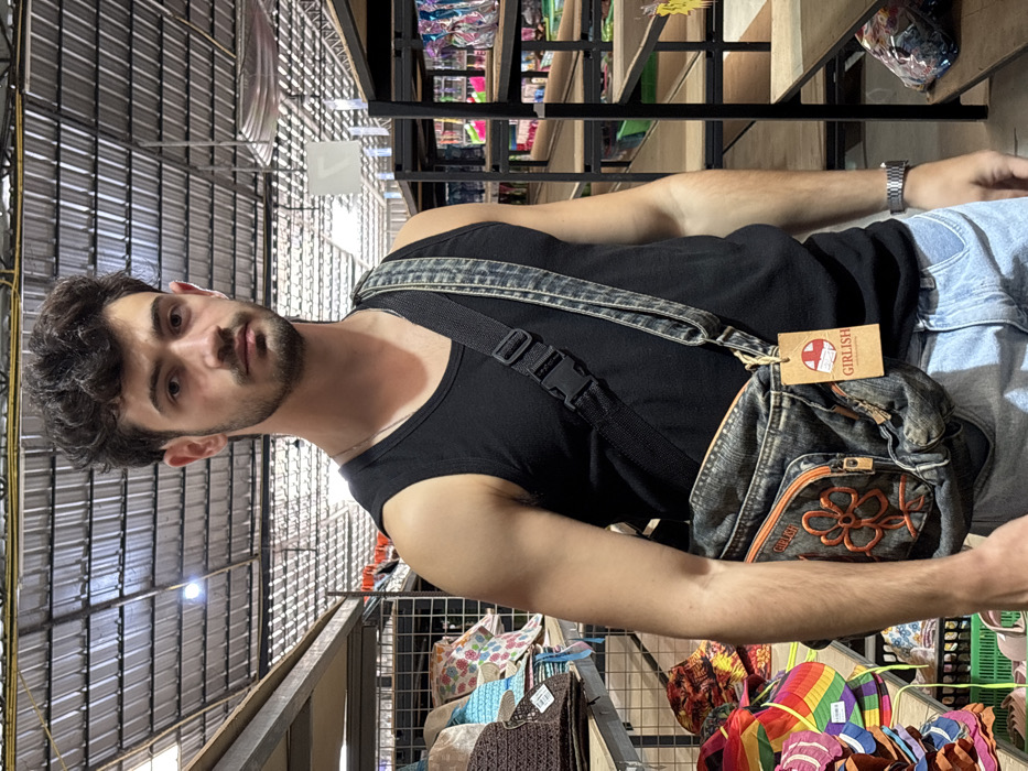A man in a black tank top and jeans stands in an indoor market, wearing a denim shoulder bag labeled "GIRLISH." The market features metal shelves packed with merchandise, including colorful items on the bottom shelf. The ceiling is made of corrugated metal. The man has dark hair and a short beard.