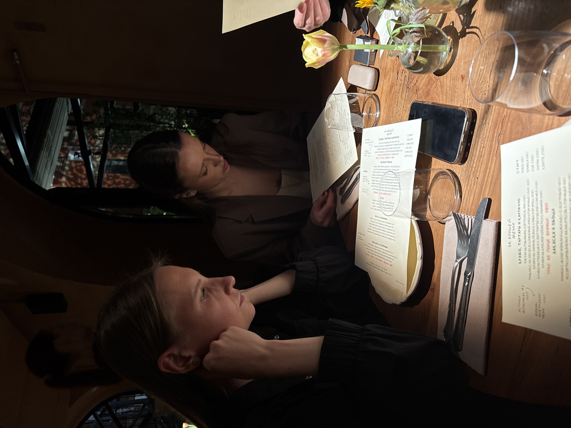 Two young women sit at a wooden table in a restaurant. They are looking at menus. A vase with a yellow tulip sits on the table with glasses, cutlery, a phone, and other objects. The restaurant appears to be in a historic building based on the architecture seen through the windows. The menus are in Russian, suggesting the location is likely in Moscow, Russia.