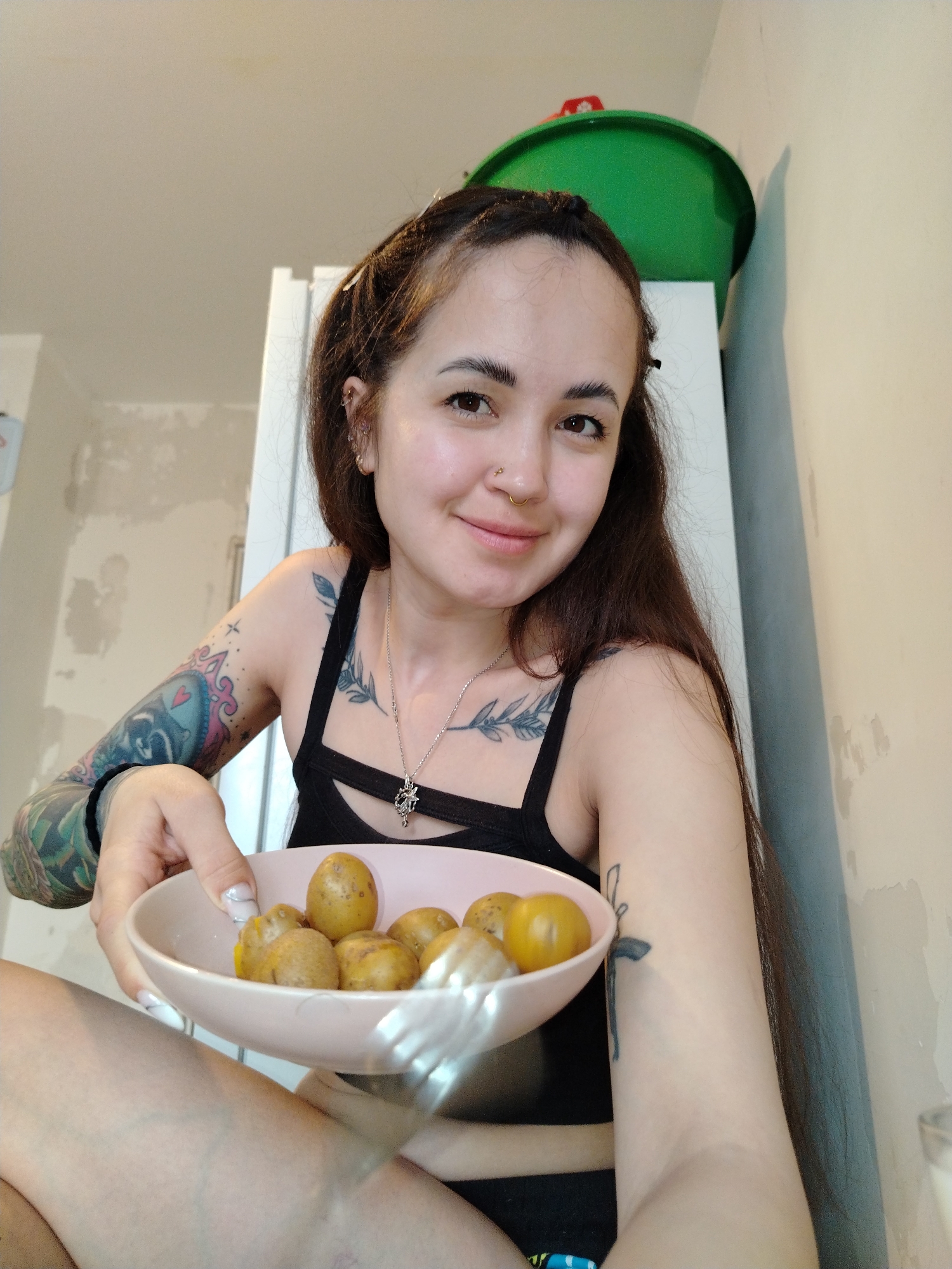 A young woman sits cross-legged in an interior space that appears to be a kitchen or living area, holding a pink bowl filled with small, cooked potatoes. She is smiling at the camera. She is wearing a black tank top, a necklace with a pendant, and black bottoms. Her left arm is heavily tattooed, and there is a smaller tattoo on her right arm. The background includes a white refrigerator-like appliance, topped with a green plastic bucket. There is exposed wall plaster or damaged paint behind the appliance. The location is Noviy, Russia.