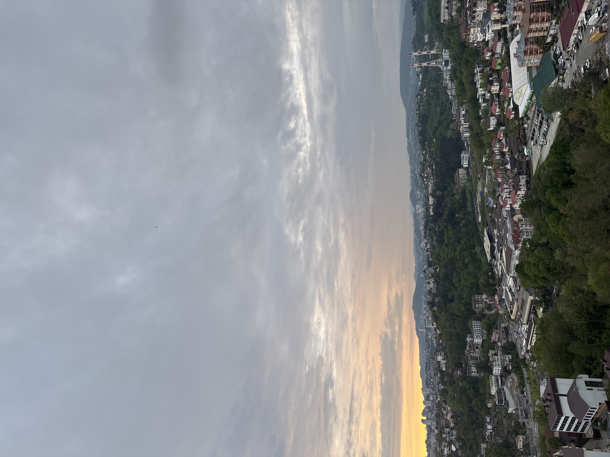 The image shows an aerial view of a town in Sochi, Russia. The foreground is the sky with clouds and a hint of a sunset glow. The town below is filled with buildings of various sizes and colors, interspersed with trees and vegetation. The buildings appear to be a mix of residential and commercial structures. A mountain range can be seen in the background, adding depth to the scene. There are some cars and roads visible in the lower right side of the image.