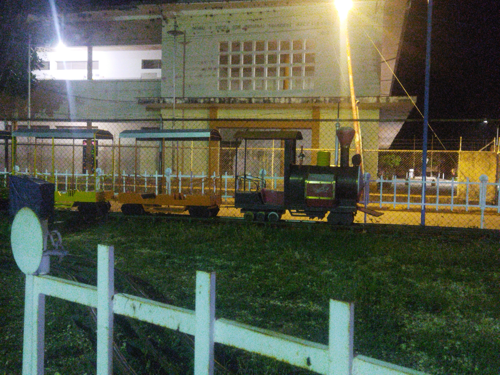 The image shows a small, colorful train with several cars parked on a grassy area next to a white fence at night. Behind the train, there's a chain-link fence, and behind that, a two-story building with a distinctive grid of windows on the second floor. A bright streetlight casts a strong glare on the scene. The image location is Unknown, Unknown.