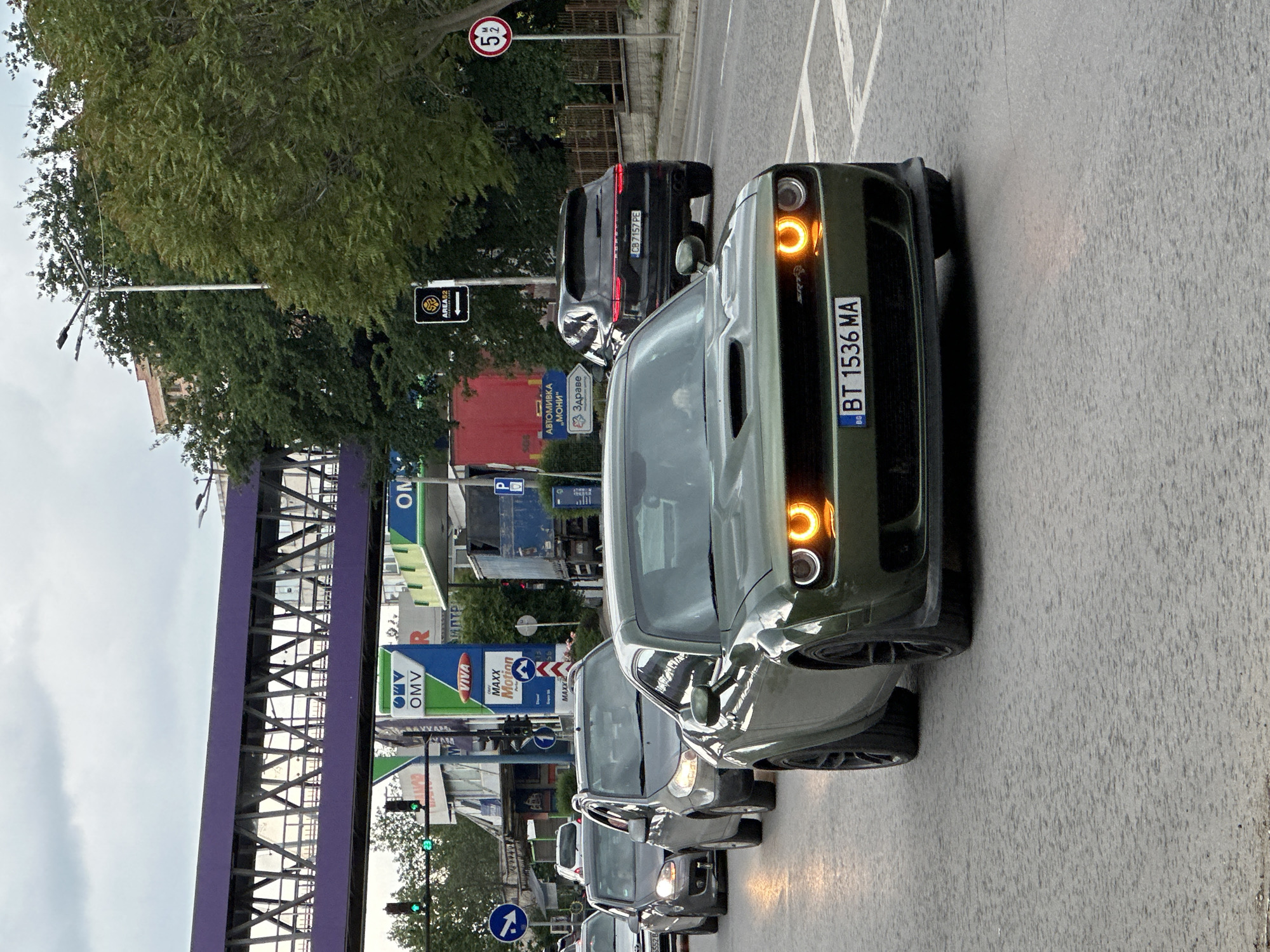 The image shows a street scene in Veliko Tarnovo, Bulgaria, likely captured during daylight hours on a cloudy day. A line of cars is visible on the road, with an olive-green Dodge Challenger prominently positioned in the foreground. The car has a license plate reading "BT 1536 MA", indicating it's registered in the Veliko Tarnovo region. Behind the Challenger, there is a black Porsche Cayenne with the license plate "CB 7157 PE". A speed limit sign with "52" (kilometers per hour) is visible, suggesting a controlled speed zone. Various commercial signs and infrastructure elements are present, including an OMV gas station and a purple overhead structure, possibly a bridge or walkway. The presence of a directional road sign, a light pole, and green traffic lights indicate the location is a relatively busy intersection or main thoroughfare.