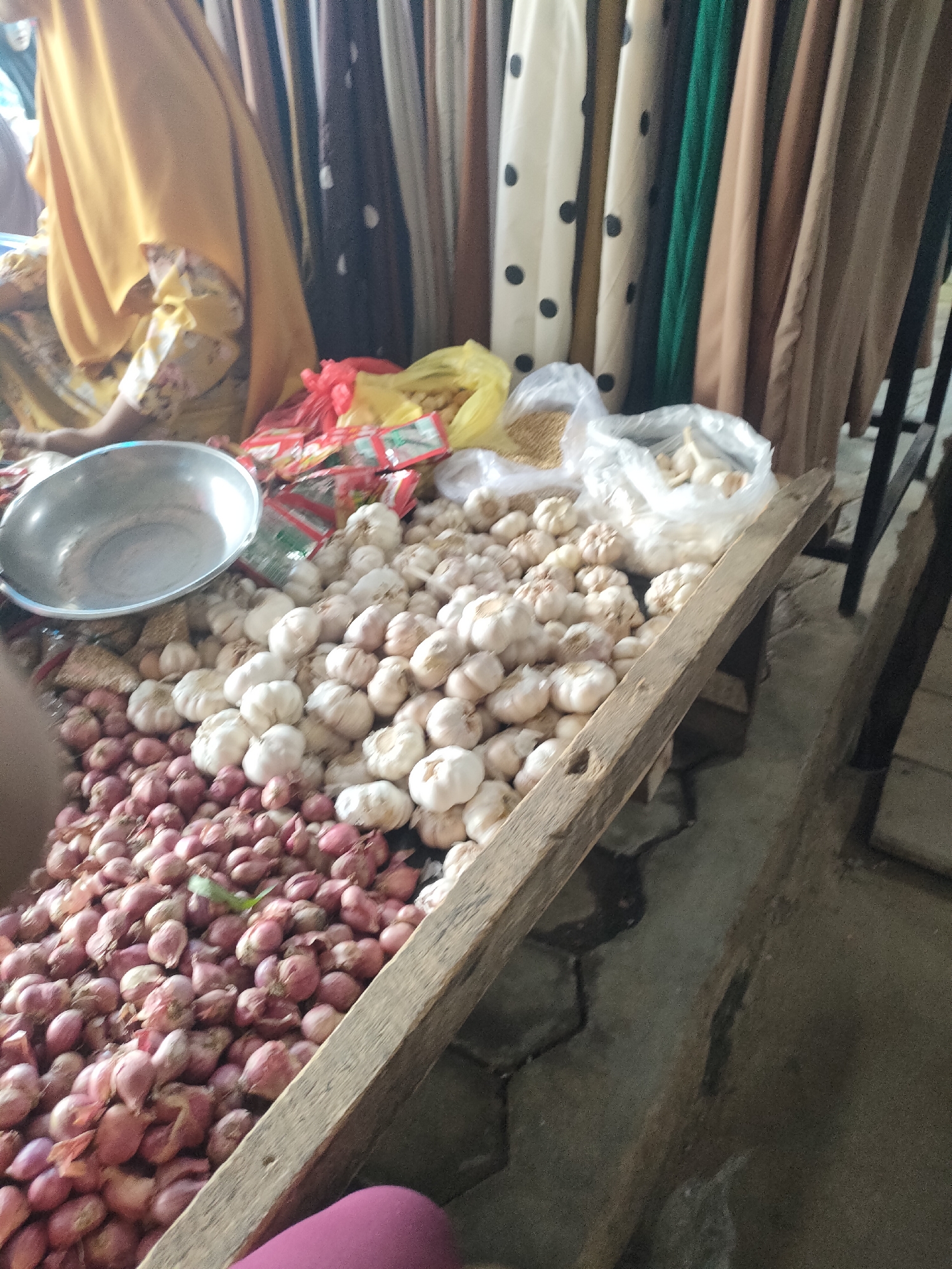 The image depicts a market stall in Sukoyoso, Indonesia, featuring a variety of produce and textiles. In the foreground, a large pile of red onions is visible, with a scattering of garlic bulbs on top. A metal bowl is placed on the onions. Behind the produce, there are several plastic bags containing more garlic and what appears to be dried grains. The background displays a collection of hanging textiles, including patterned fabrics and solid-colored drapes. A person, partially visible and wearing a yellow headscarf and patterned clothing, is seated at the stall, suggesting engagement in commerce. The stall itself is constructed from rough wooden planks. The ground beneath the stall appears to be a paved surface, possibly concrete tiles. The overall impression is one of a bustling local market environment.