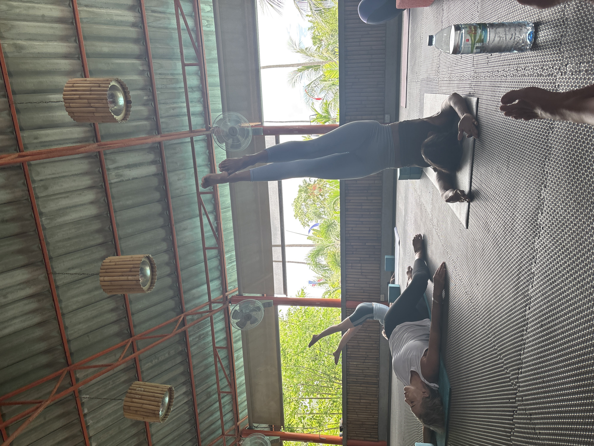 This image shows an indoor yoga class taking place in Rawai, Thailand. Two women are visible on yoga mats, engaged in different poses on a dark grey mat floor. A water bottle is on the mat. The room has a red steel frame supporting the roof, with round bamboo lamps hanging from the ceiling. Outside, trees and greenery are visible through open walls.