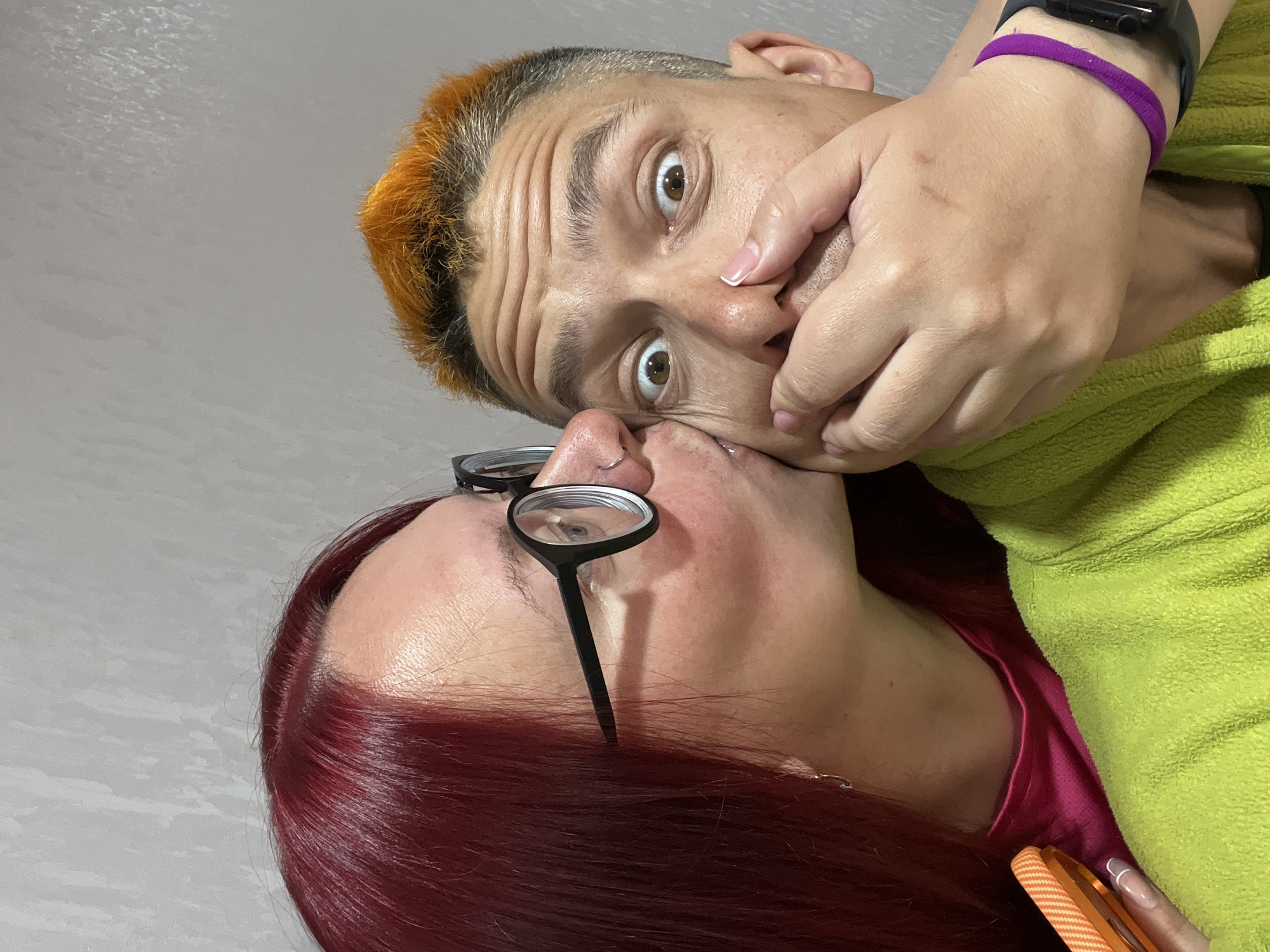 The image shows two individuals in an intimate moment. One person, with short hair dyed orange at the front, has their mouth covered by the hand of another person wearing a green sweatshirt. The person being touched has wide eyes and a surprised expression. The second person, with long, red hair and glasses, is close to the first person, appearing to be about to kiss them on the nose. The scene is indoors and the background is a plain, light grey color. There is no information available to determine the location as the city of Chaikovsky, Russia.
