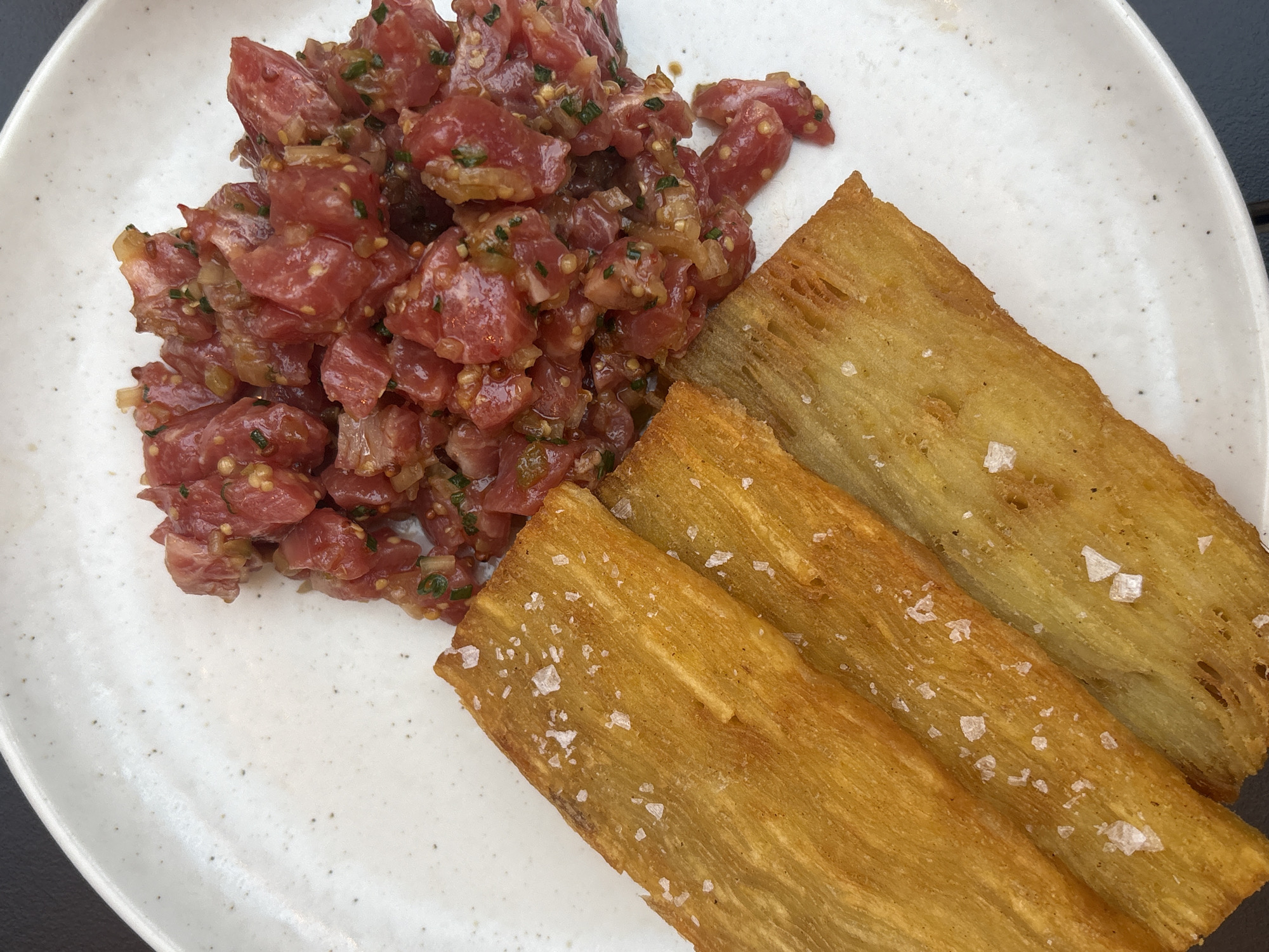 The image shows a white speckled plate with a serving of diced raw meat, likely tuna tartare, mixed with herbs and other ingredients. Next to the meat are three rectangular, golden-brown items, possibly potato or cassava fries, sprinkled with coarse salt. The background is a neutral, possibly a table surface. It cannot be determined from the image if this location is in the city of London, United Kingdom.