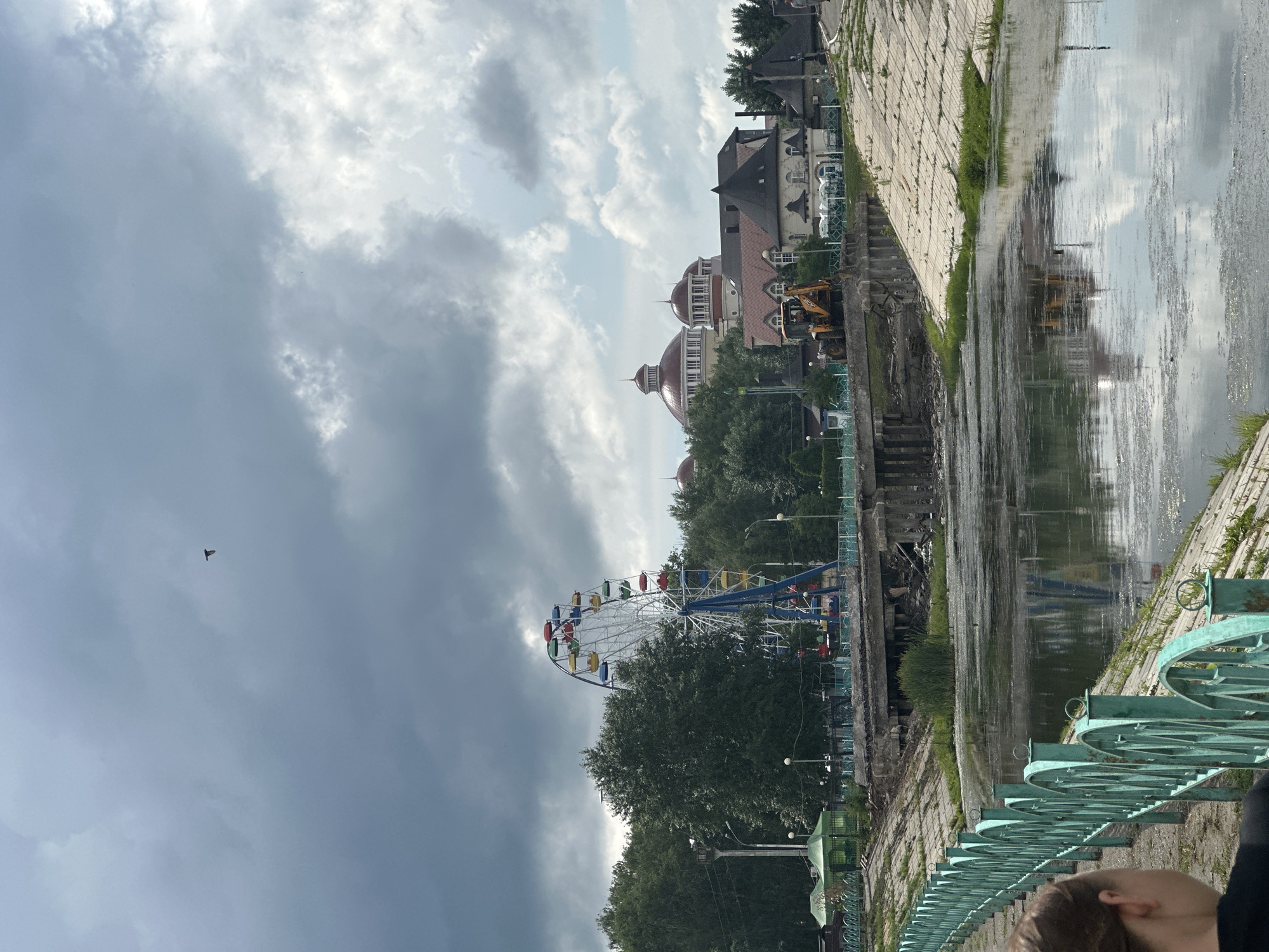 The image depicts an outdoor scene in Saransk, Russia, under a cloudy sky. A body of water, possibly a river or canal, occupies the foreground and reflects the sky and surrounding structures. A green metal fence runs along the edge of the water. In the background, a Ferris wheel with colorful cabins is visible amidst a dense green tree line. Behind the trees, several buildings are present, including structures with domed roofs and a yellow excavator working near a damaged bridge. The overall atmosphere suggests an urban park or recreational area undergoing some form of construction or renovation.