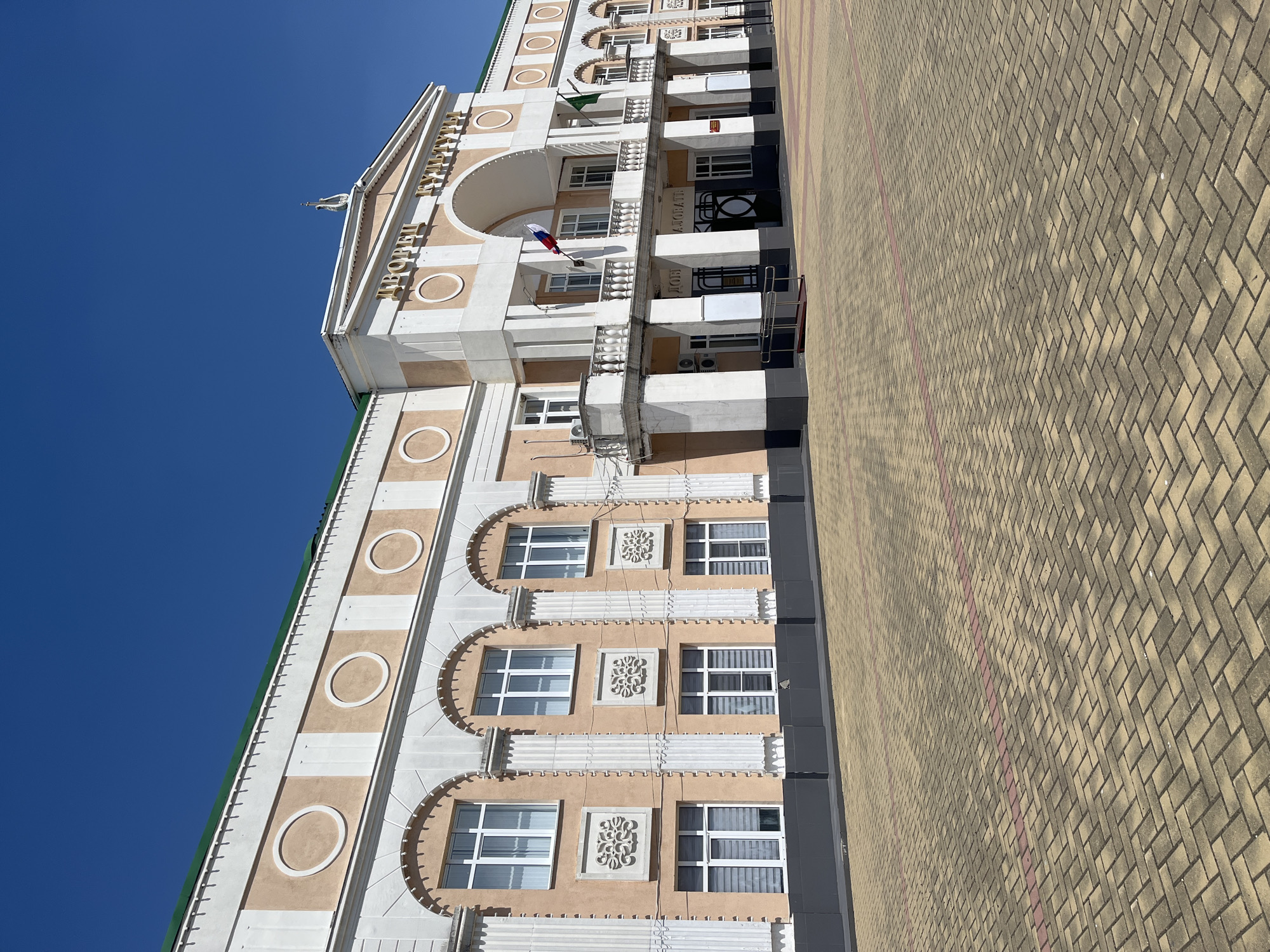 The image shows a wide, eye-level, outdoor, daytime shot of the Palace of Culture in Takhtamukai, Russia. The building is a large, light-peach colored structure with white architectural details, including columns, arches, and circular decorative elements on the upper facade. The words "ДВОРЕЦ КУЛЬТУРЫ" (Palace of Culture) are visible in gold lettering above a central arched entrance. A Russian flag and a green flag are displayed on balconies. The building is set back from a paved courtyard made of tan and red interlocking bricks arranged in a herringbone pattern. The sky above is clear and bright blue. There are no people or animals visible in the image.