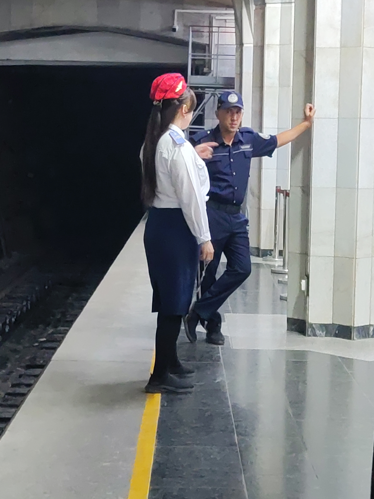 In Tashkent, Uzbekistan, two individuals are standing on a subway platform. On the left is a woman wearing a red and gold hat, a white collared shirt, and a dark blue skirt. She has long dark hair. On the right is a man in a dark blue uniform with a matching cap. He is leaning against a white tiled pillar and appears to be speaking with the woman. The platform has a yellow safety line along the edge closest to the tracks. The background shows a dark tunnel and large tiled pillars.