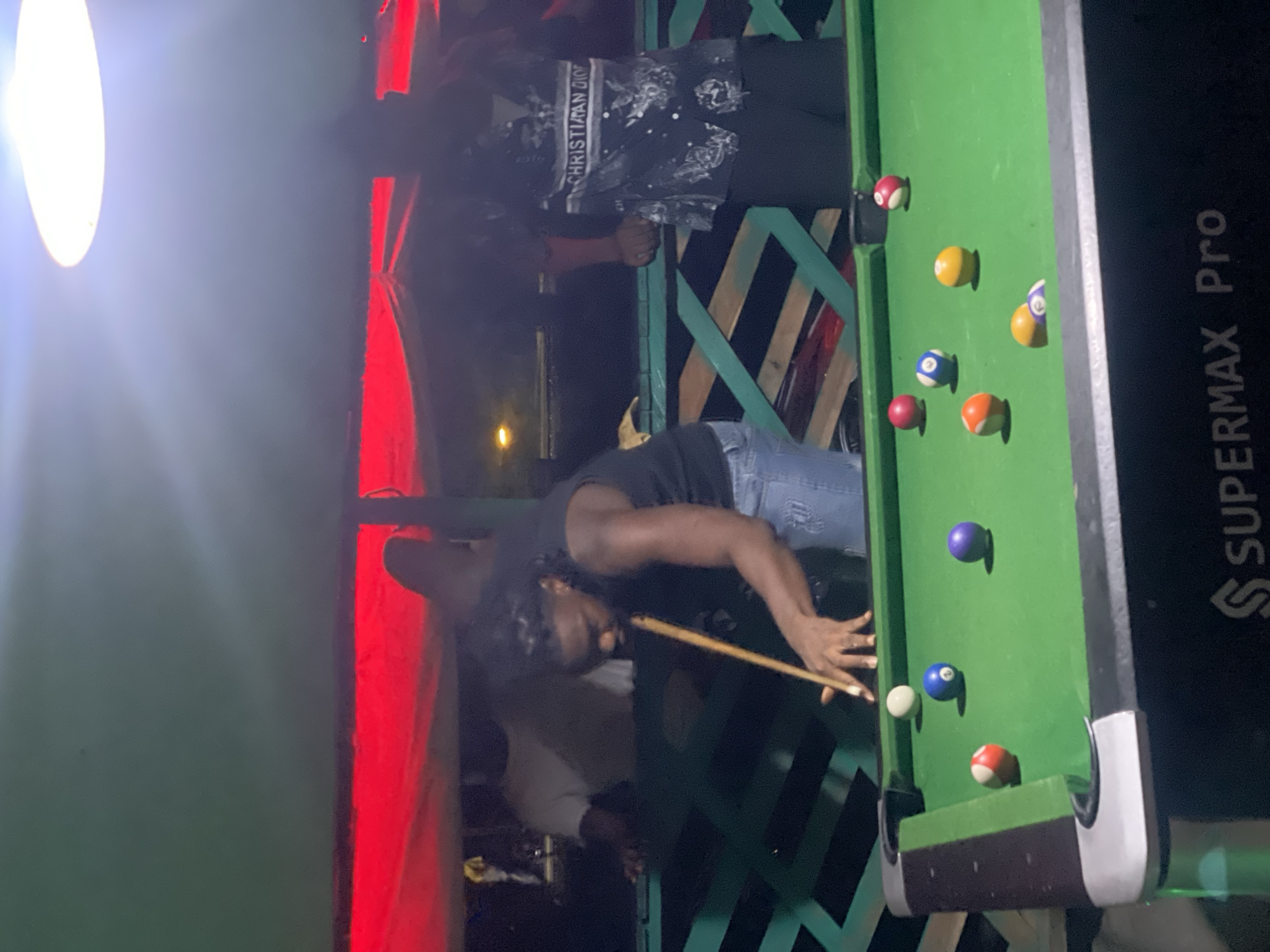 A person is playing pool on a "SUPERMAX Pro" pool table. The table is green with several pool balls on it. Another individual stands nearby. The scene is set in what appears to be a dimly lit outdoor area in Lokoja, Nigeria. The lighting is harsh with a bright light source nearby. A red vertical structure is visible on the left side of the image. There are green wood slats in the background. The person playing pool is wearing a dark shirt and jeans, and holding a pool cue. The other individual is wearing a Christian Dior shirt.