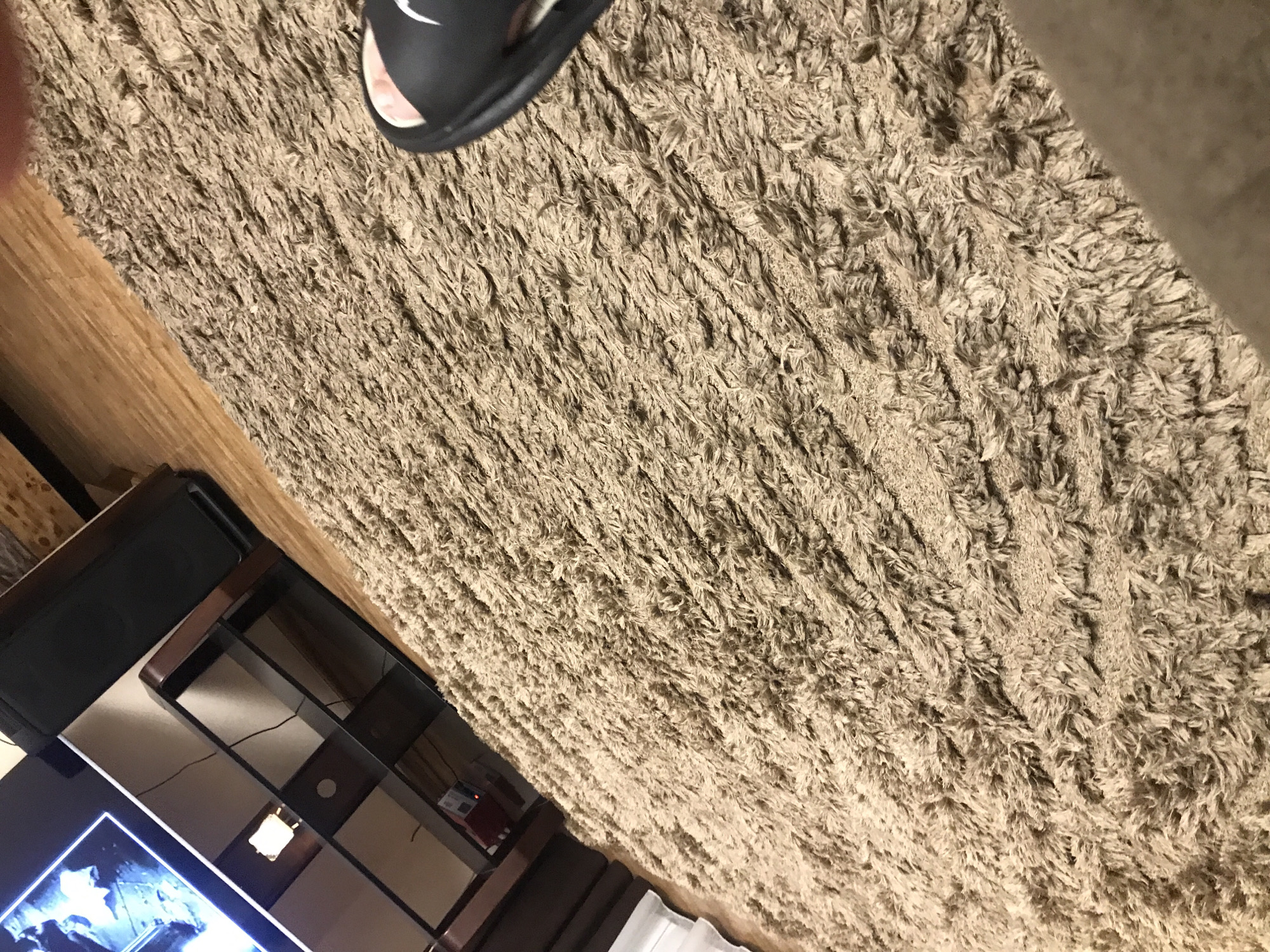The image depicts a close-up view of a tan shag carpet with a black sandal partially visible in the upper portion of the frame. A television, shelves, and an adjacent section of wooden flooring can be seen in the lower left quadrant of the image. The composition suggests an indoor setting, likely a living room. The city of Yerevan, Armenia, is not discernible from the details provided in this image.
