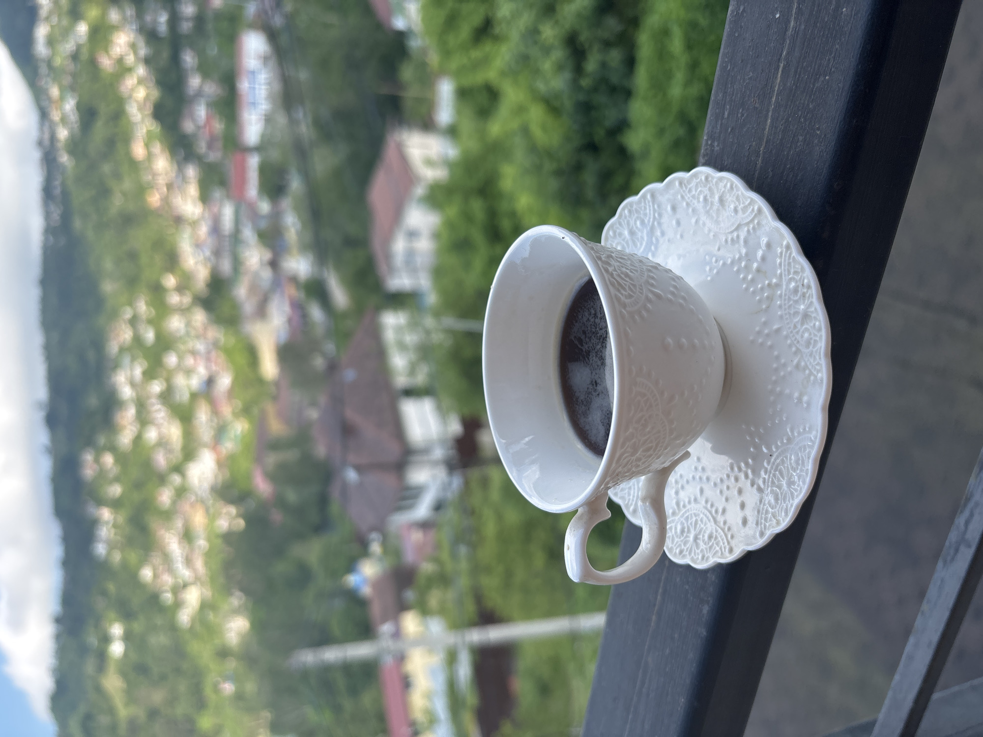 A white ceramic coffee cup and saucer with an embossed floral pattern sit on a dark wooden railing. The cup is filled with dark coffee. In the background, a blurred cityscape of Sochi, Russia, is visible on a hillside. The city features numerous houses with red and white roofs nestled among lush green trees. The sky above is partly cloudy.